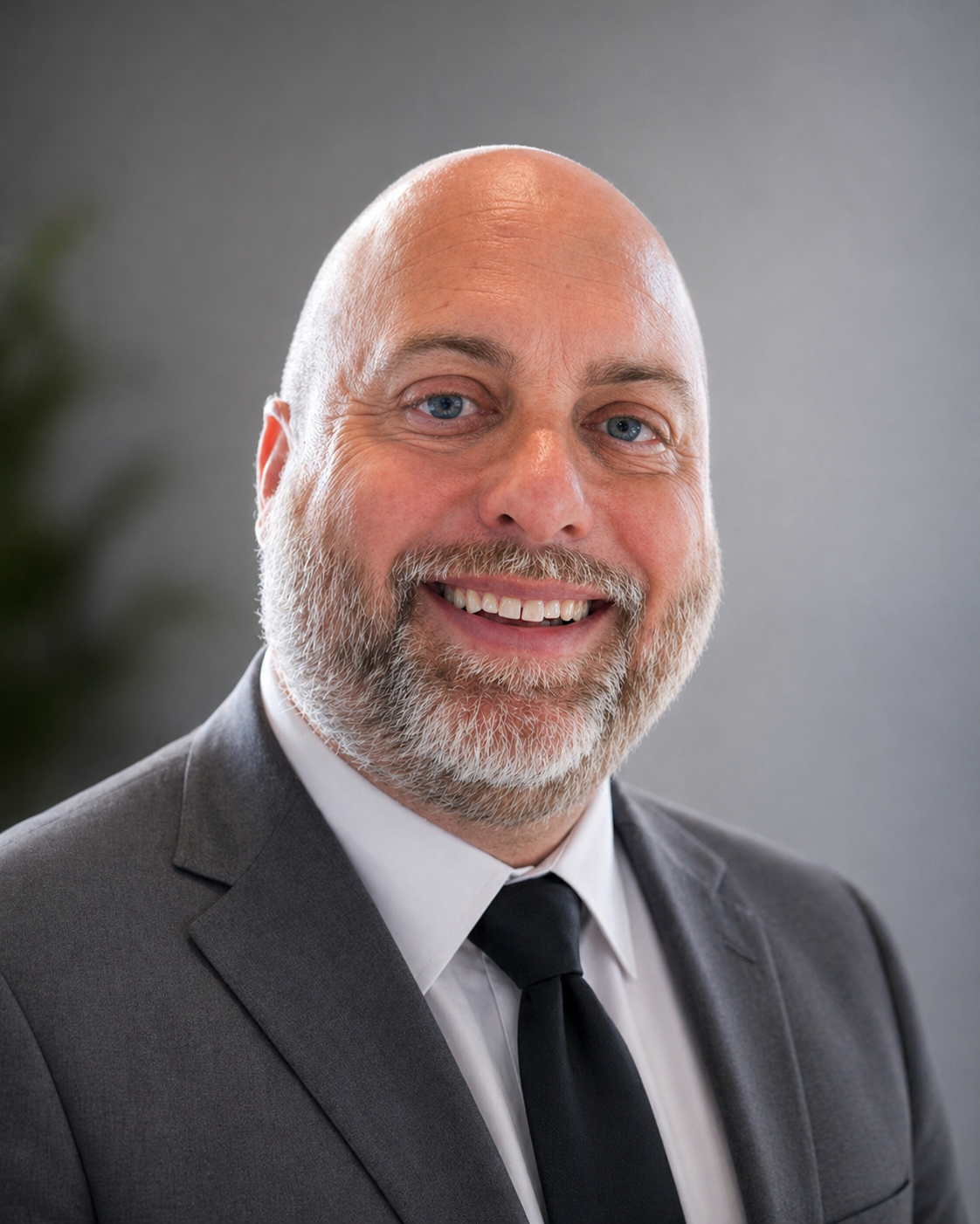 Headshot of a smiling middle-aged man with a beard, wearing a gray suit, white shirt, and black tie, against a gray background.