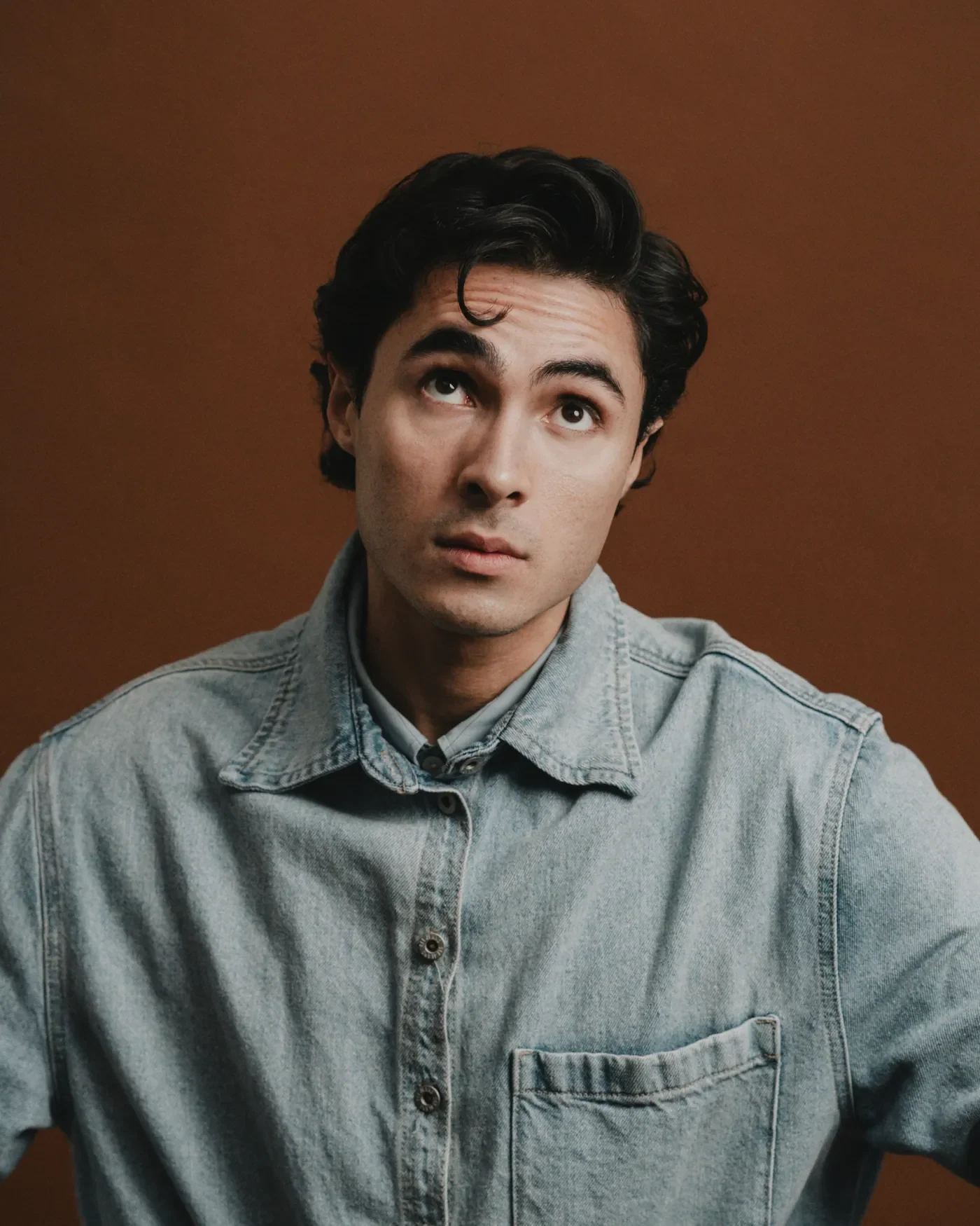 Luis del Toro actor portrait looking up wearing denim shirt against brown background