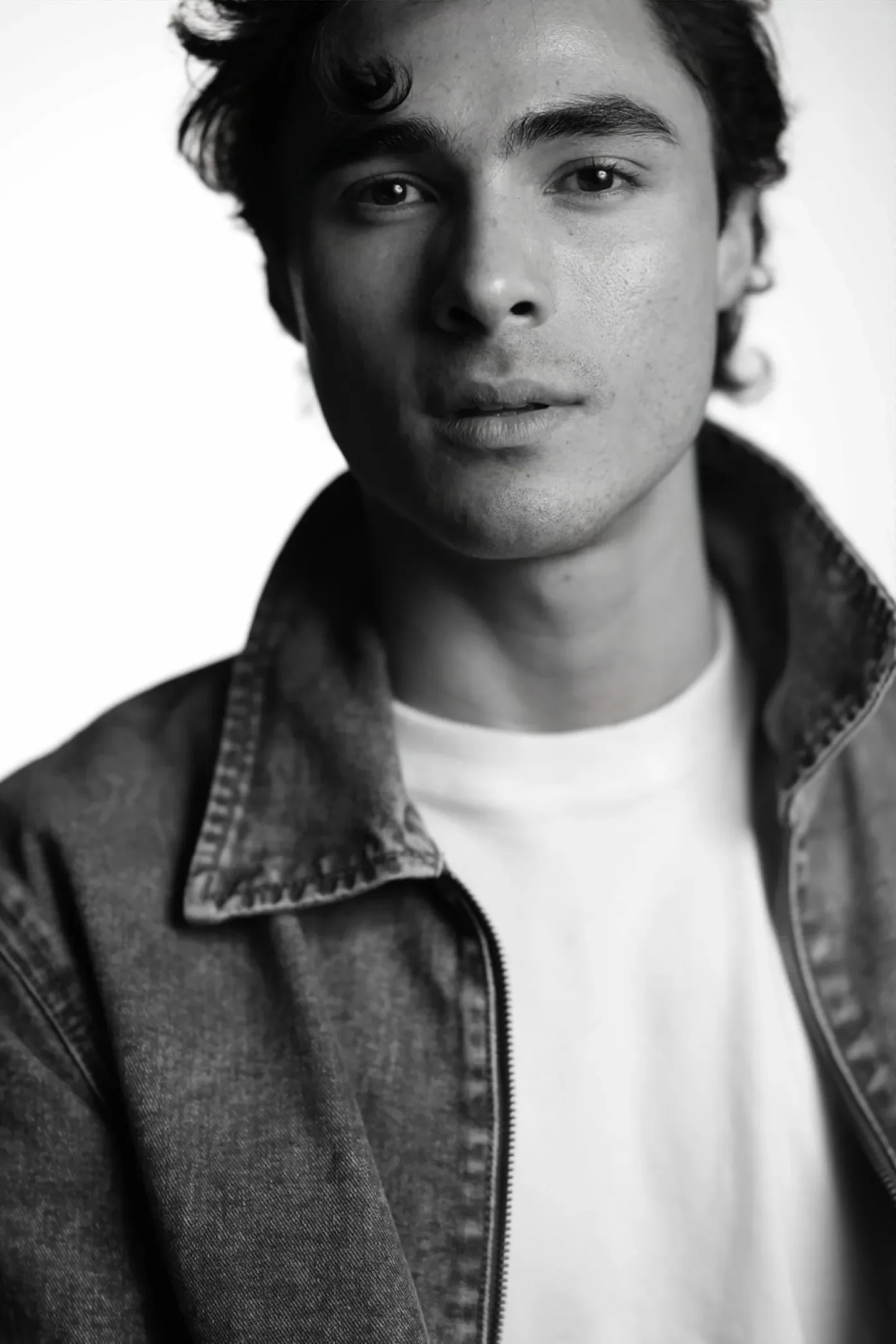 Black and white portrait of a young man with wavy hair, wearing a denim jacket over a white shirt, looking directly at the camera.