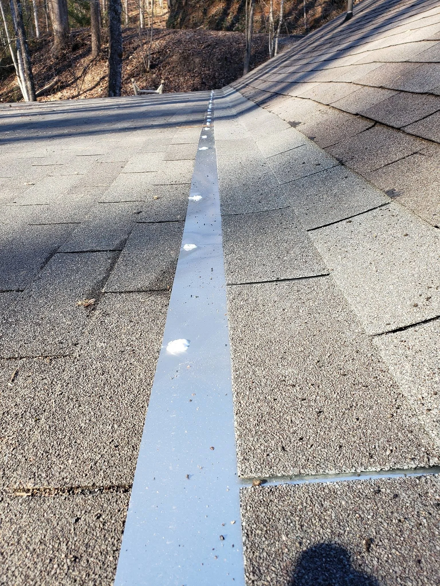The image shows a rooftop with asphalt shingles and a metal flashing strip in the center. There are some patches of snow on the roof, and the background features a wooded area with trees and a hillside.