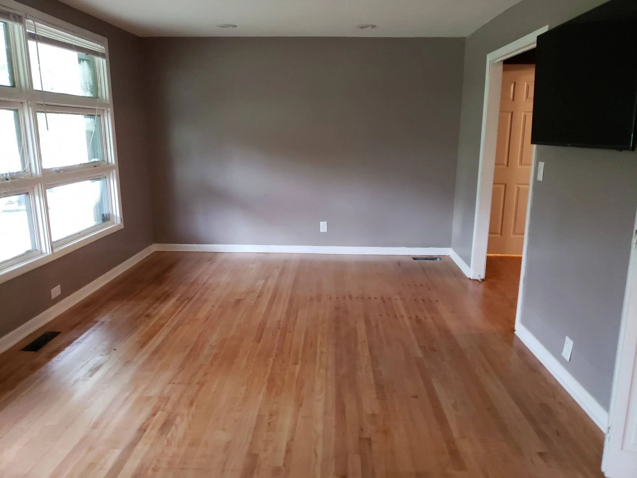 Empty living room with hardwood flooring, gray walls, large window with blinds, white baseboards, and a wall-mounted TV.