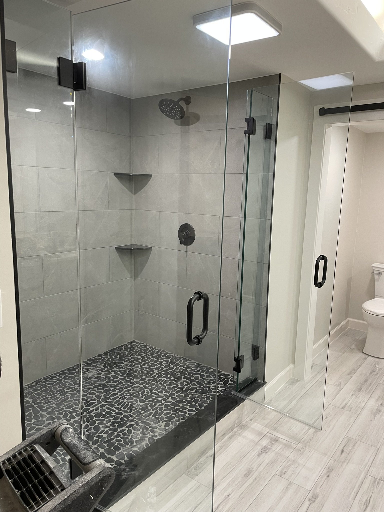 Modern bathroom with a glass-enclosed shower featuring pebble tile flooring, gray tiled walls, and a rain showerhead, with a toilet visible in the background.
