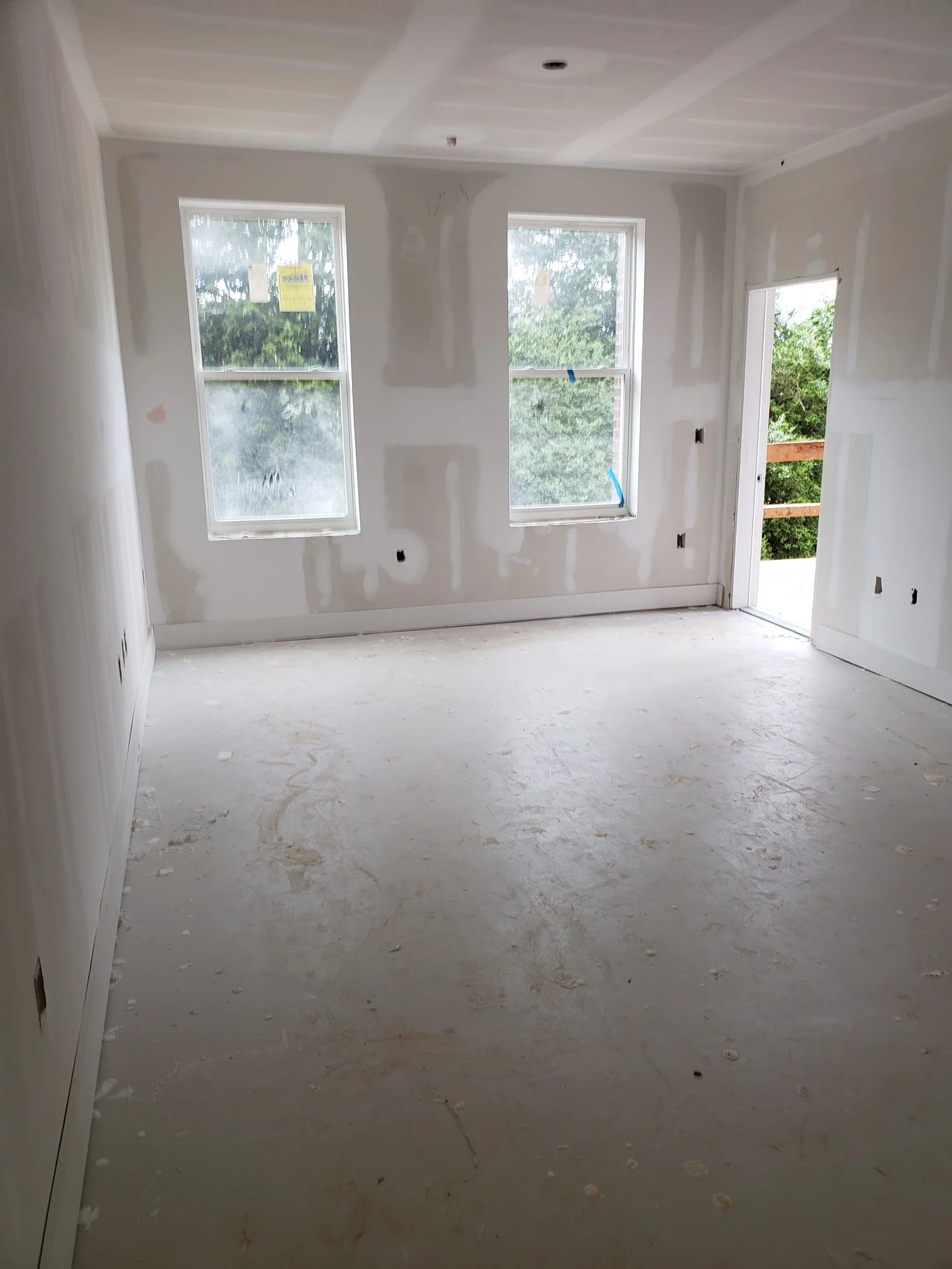 An unfinished room under construction with drywall on the walls, two windows, and an open door showing a balcony with a wooden railing. The floor is bare concrete with construction debris.