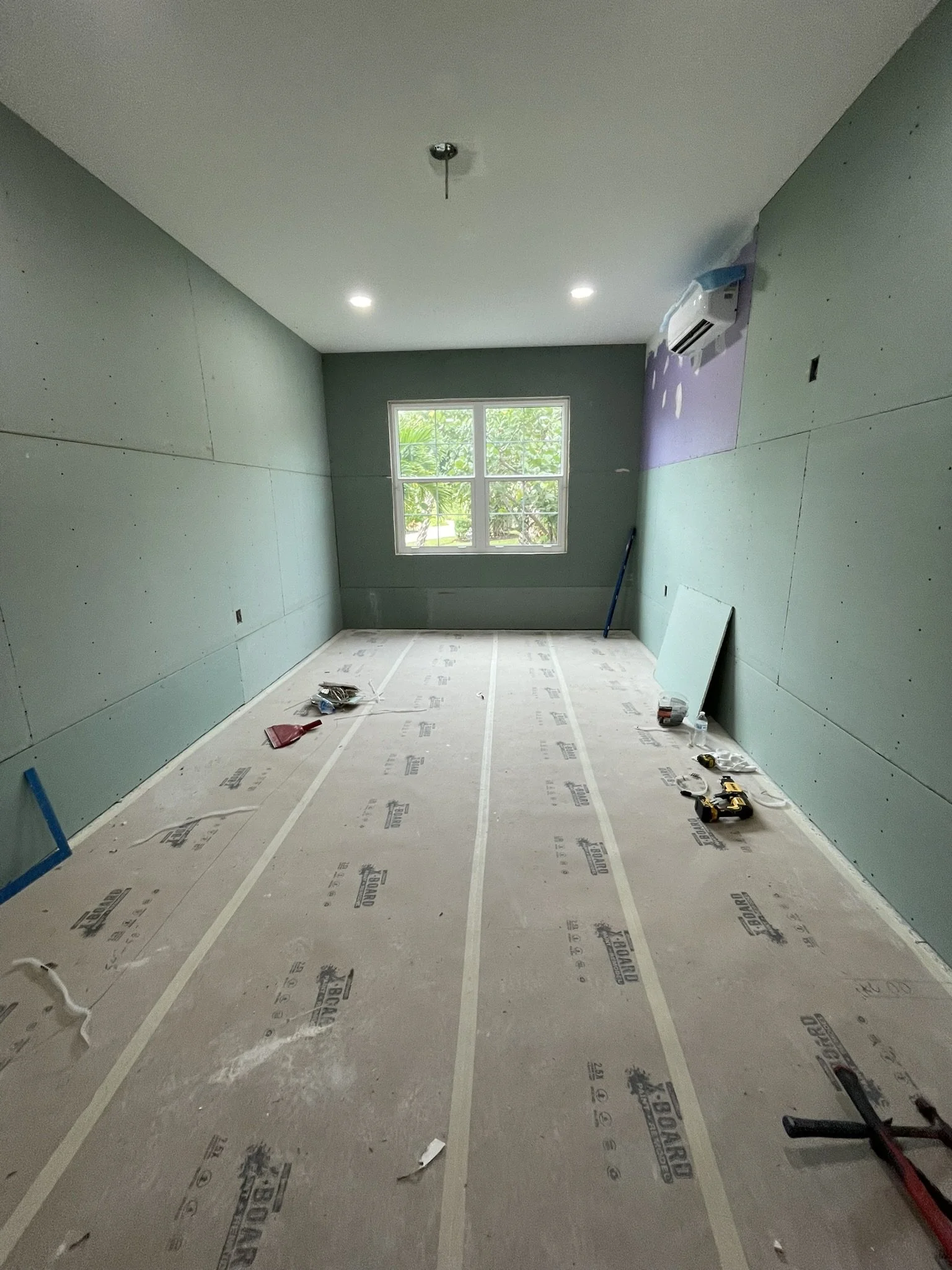 An unfinished room under construction with green drywall walls, a window, construction tools, and supplies on the floor.
