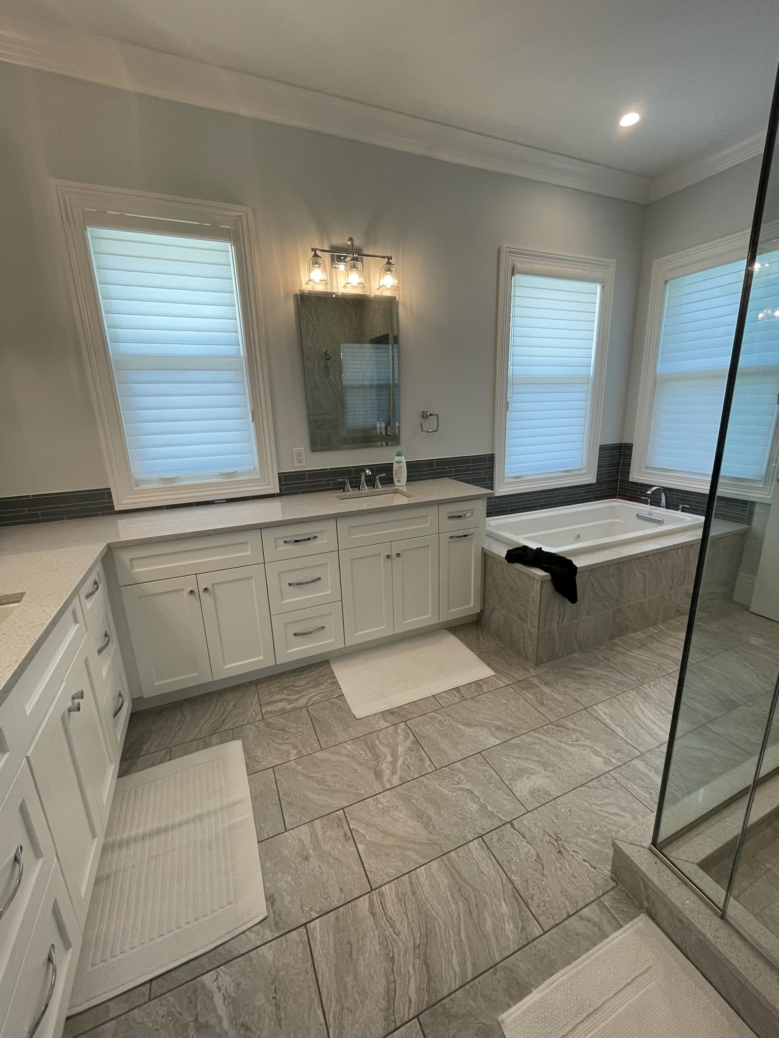 A modern bathroom with white cabinetry, a large bathtub, a glass shower, three windows with blinds, and a gray tiled floor.