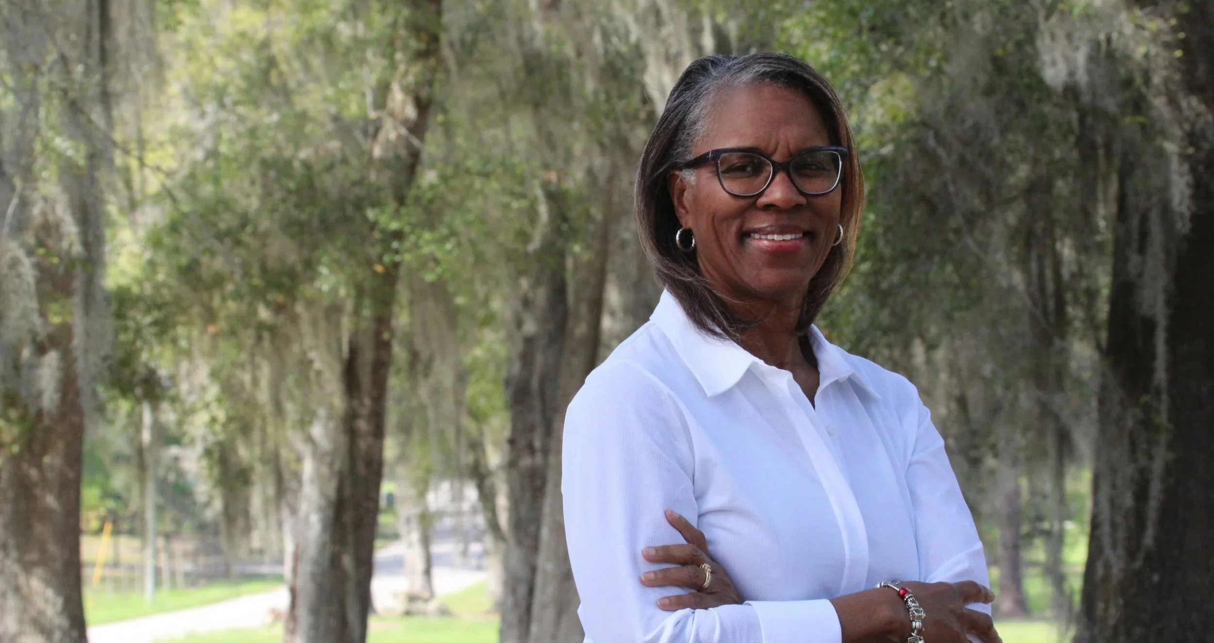 A photograph of Tina Certain with glasses and earrings, smiling with crossed arms, wearing a white shirt and black pants, standing outdoors.