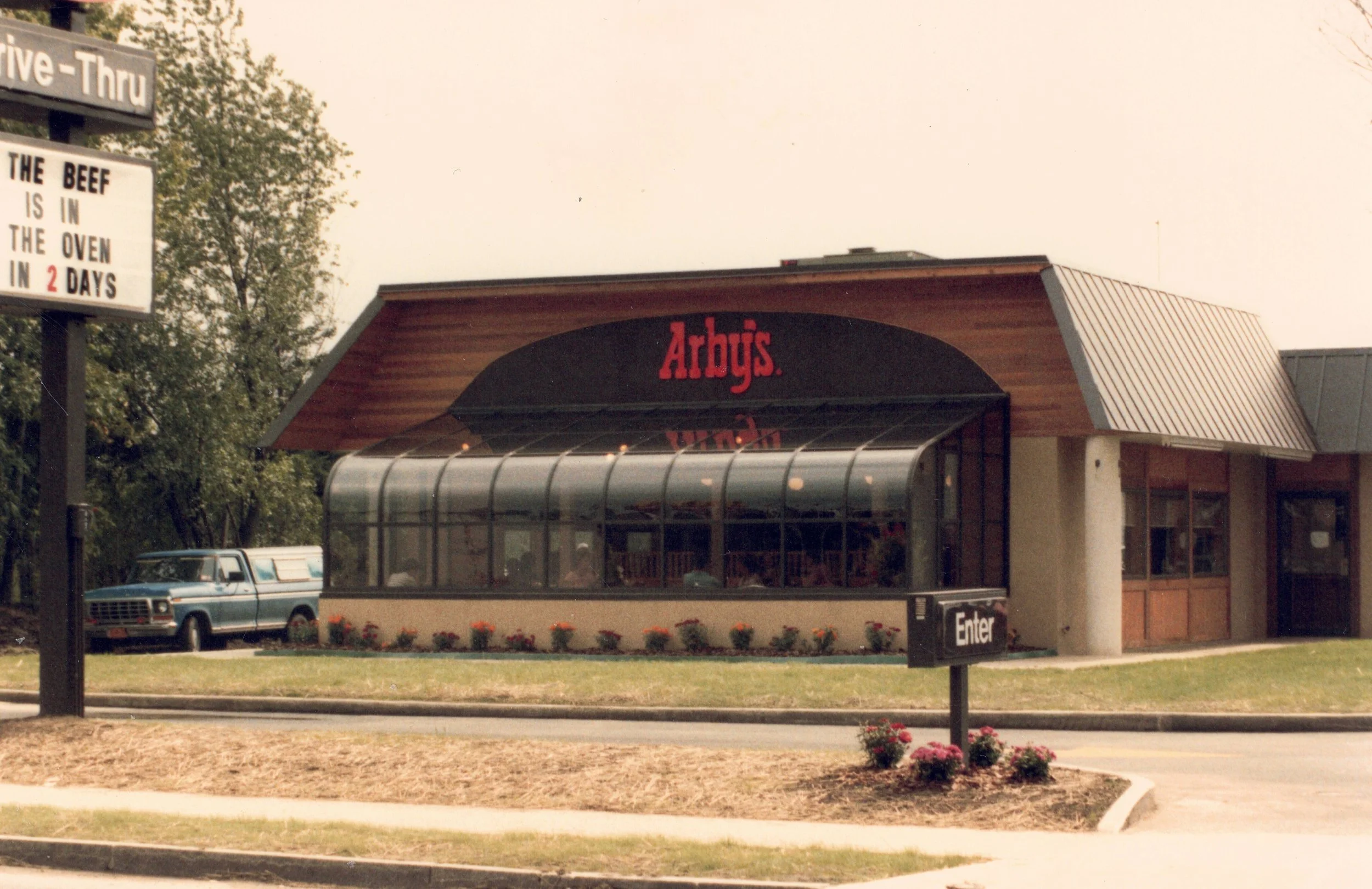 Arby's fast-food restaurant with a wooden and glass exterior, a sign that reads 'Enter', and a parking lot with a vintage car parked nearby.