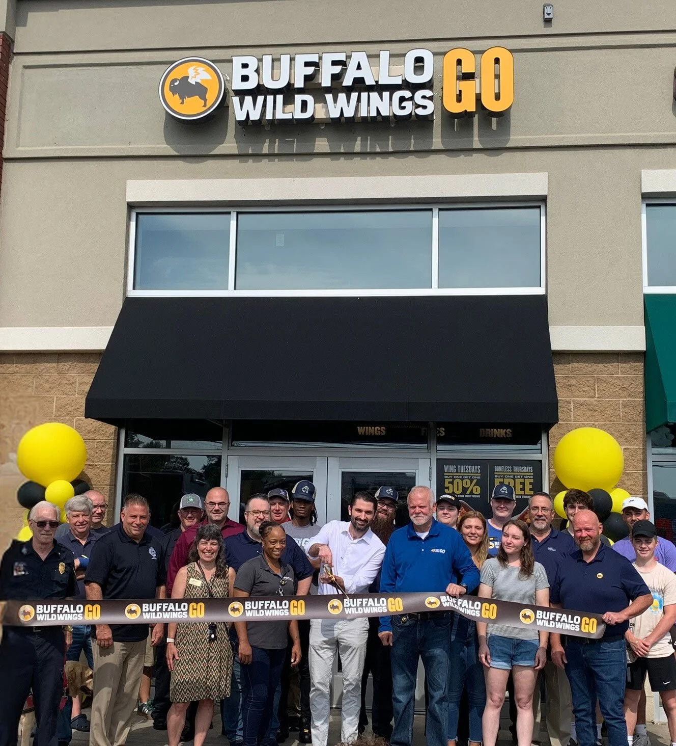 Group of people gathered for a ribbon-cutting ceremony in front of a Buffalo Wild Wings restaurant, with yellow and black balloons on either side of the entrance.