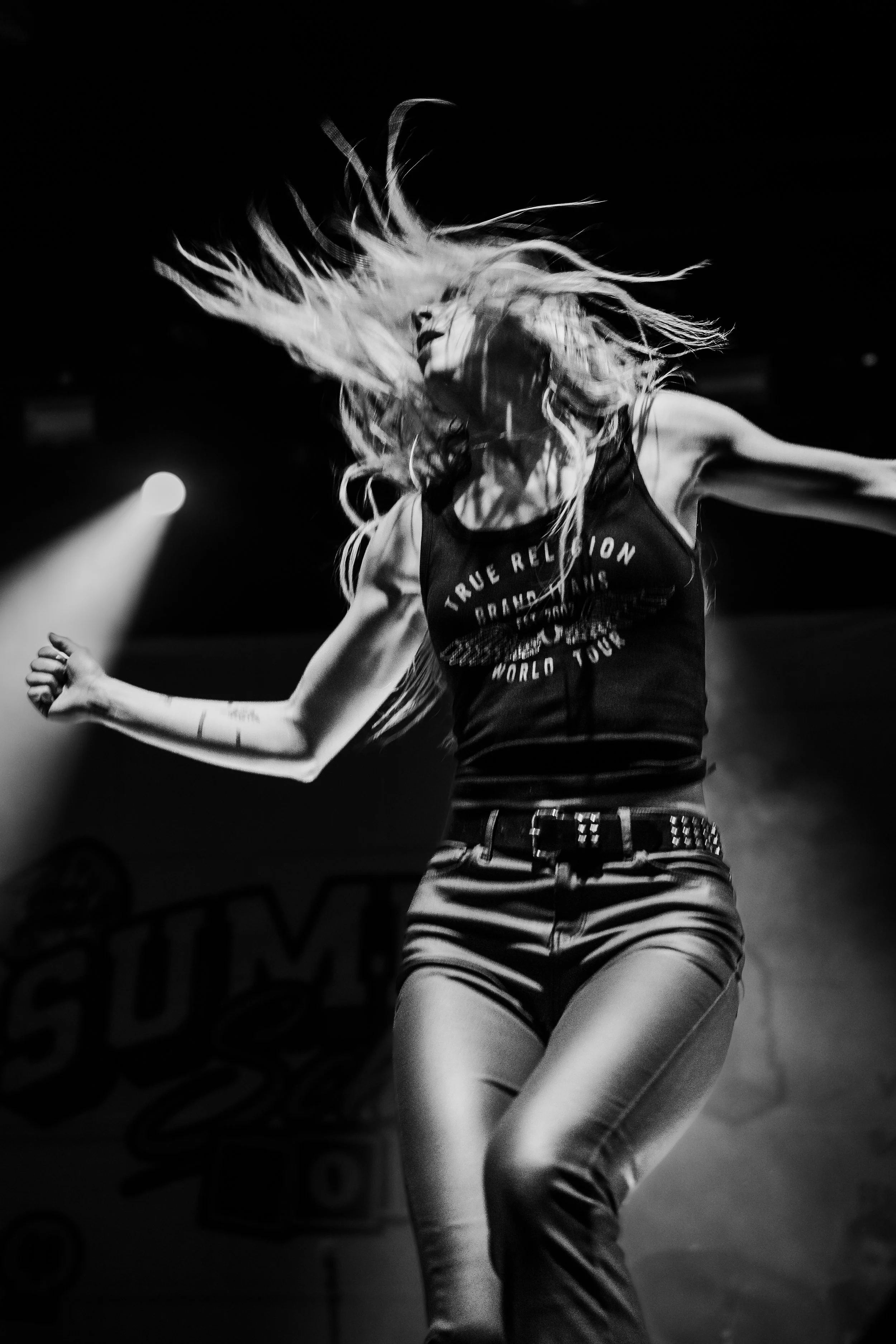 A woman with long hair dancing or jumping on stage, illuminated by a spotlight, in a black and white photo.