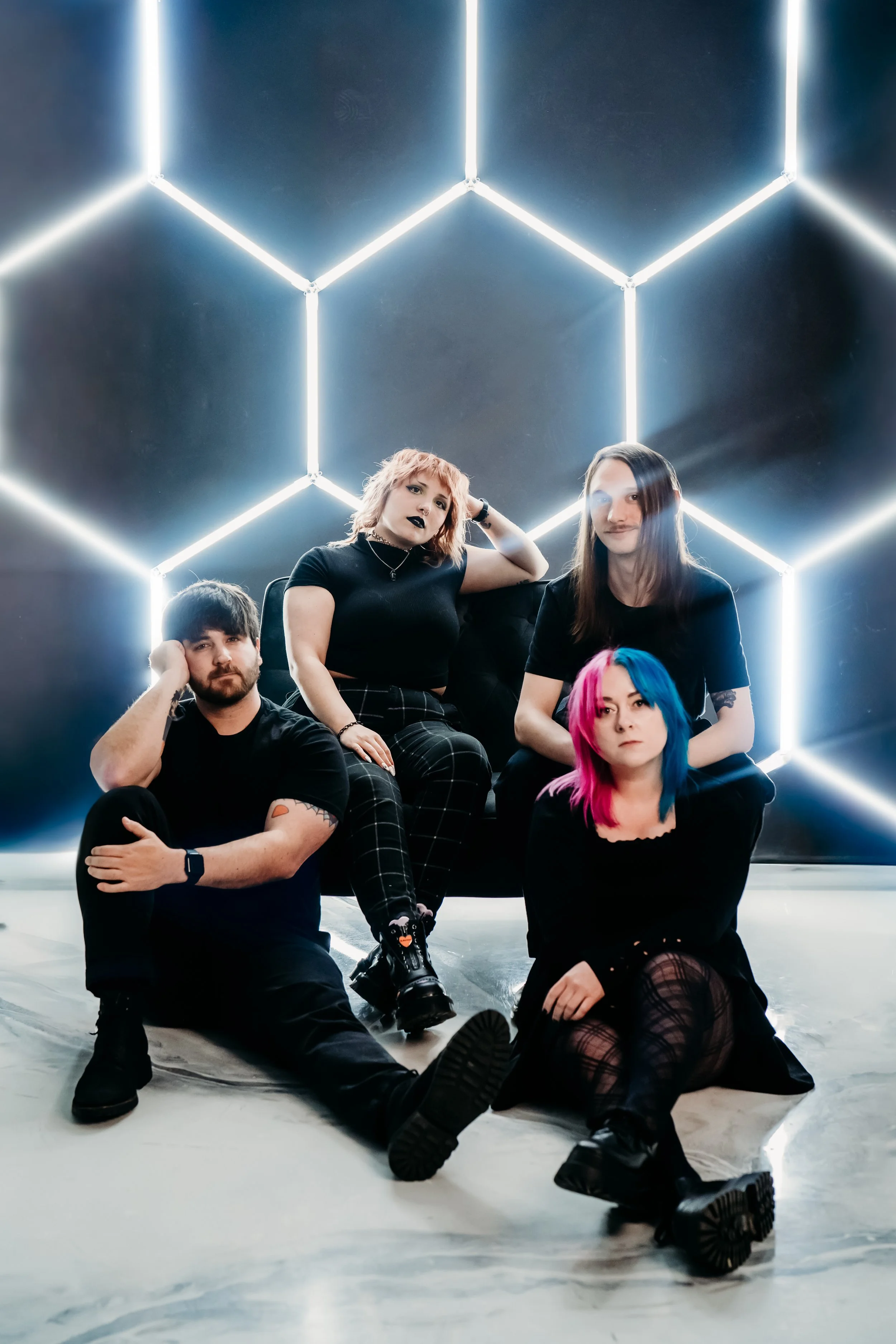 A group of four young people sitting and posing in front of a black wall with illuminated geometric shapes.