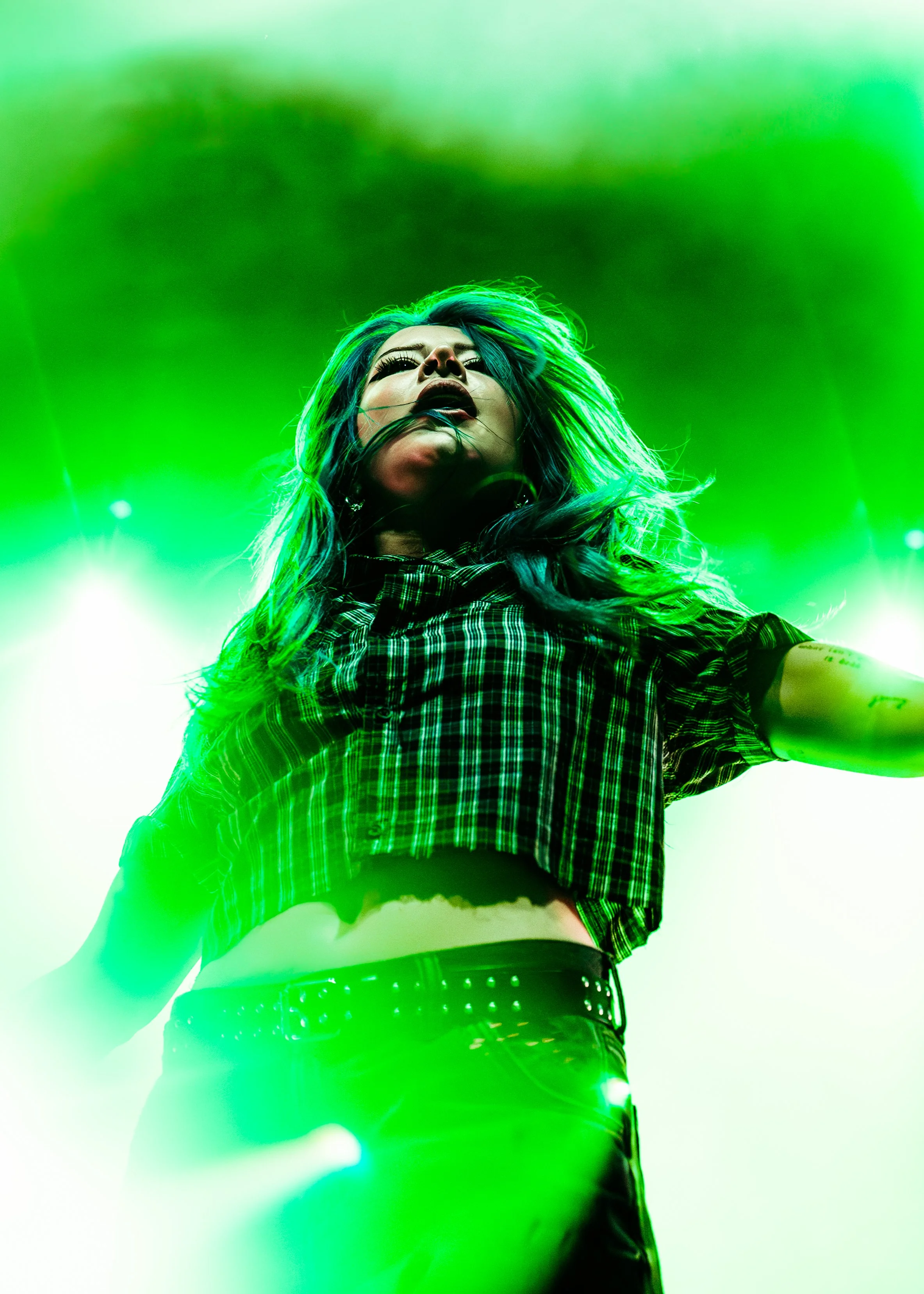 A female performer on stage illuminated by green stage lights, wearing a plaid shirt and black pants with a studded belt, with long hair and makeup, looking upward.