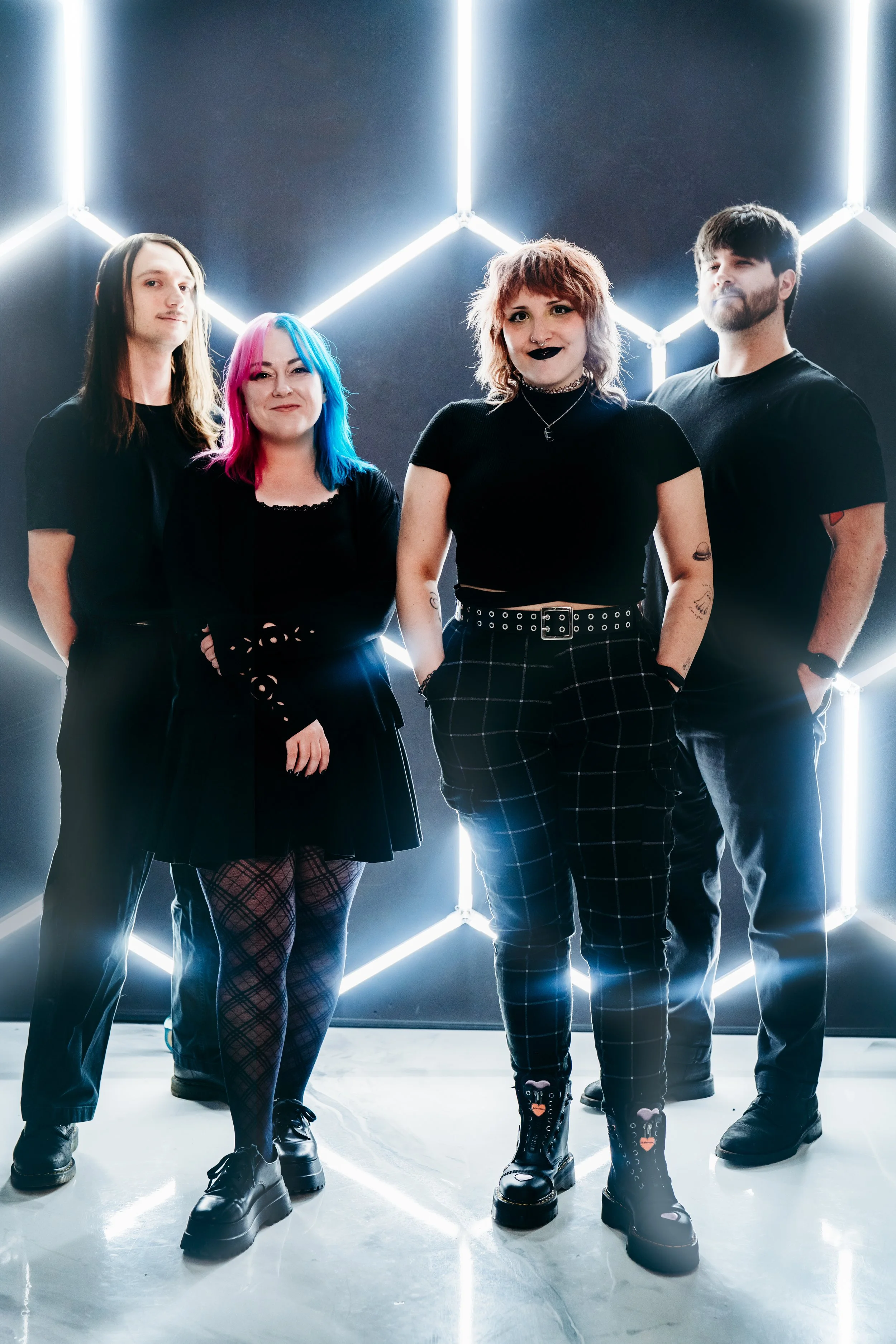 Four young people standing together in front of a geometric neon light installation, dressed in goth and alternative fashion.
