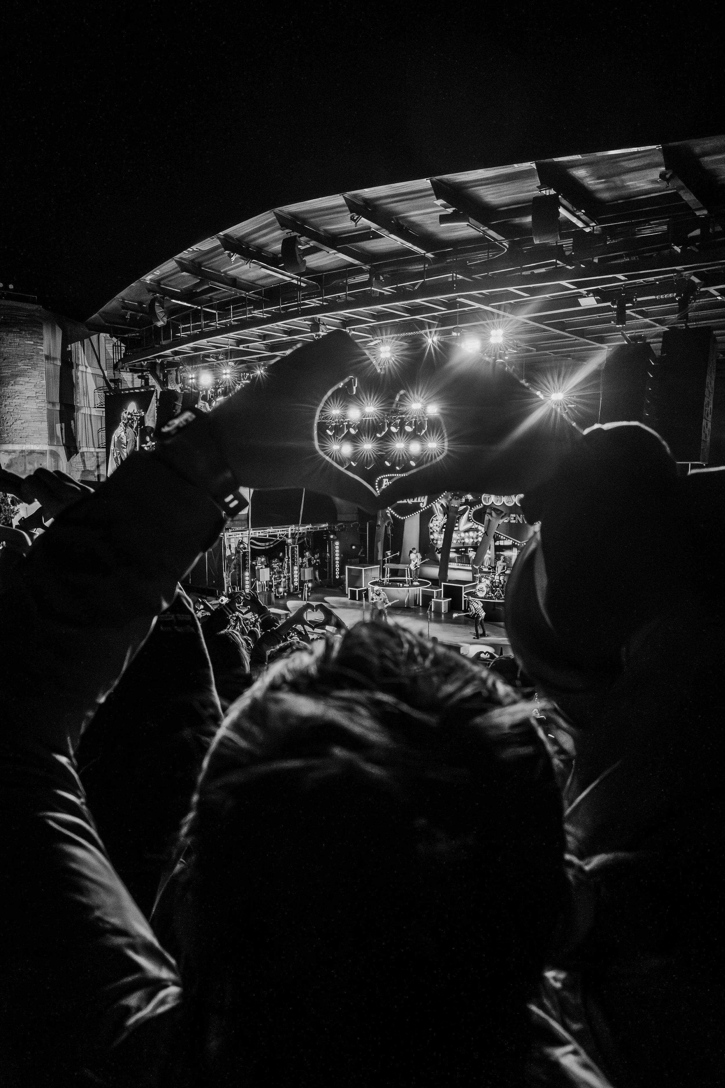A black and white photo of a concert scene with an audience making a heart shape with their hands, with a band performing on stage in the background under bright lights.
