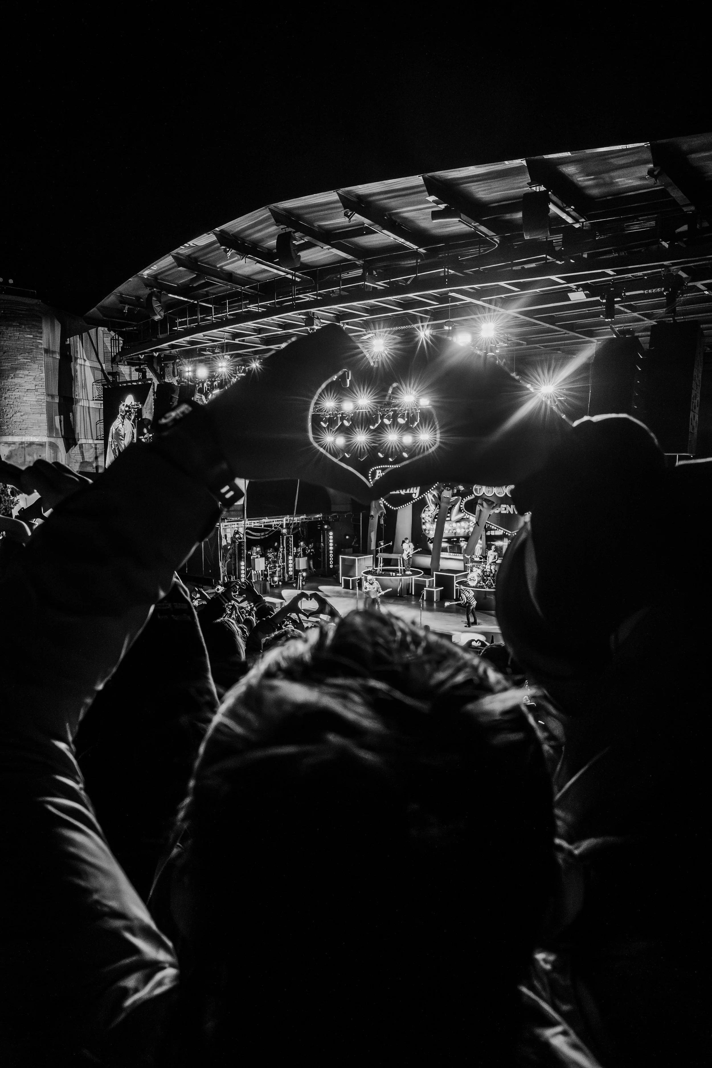 Black and white photo of a concert stage with performers and audience, with a person in the foreground making a heart shape with their hands.