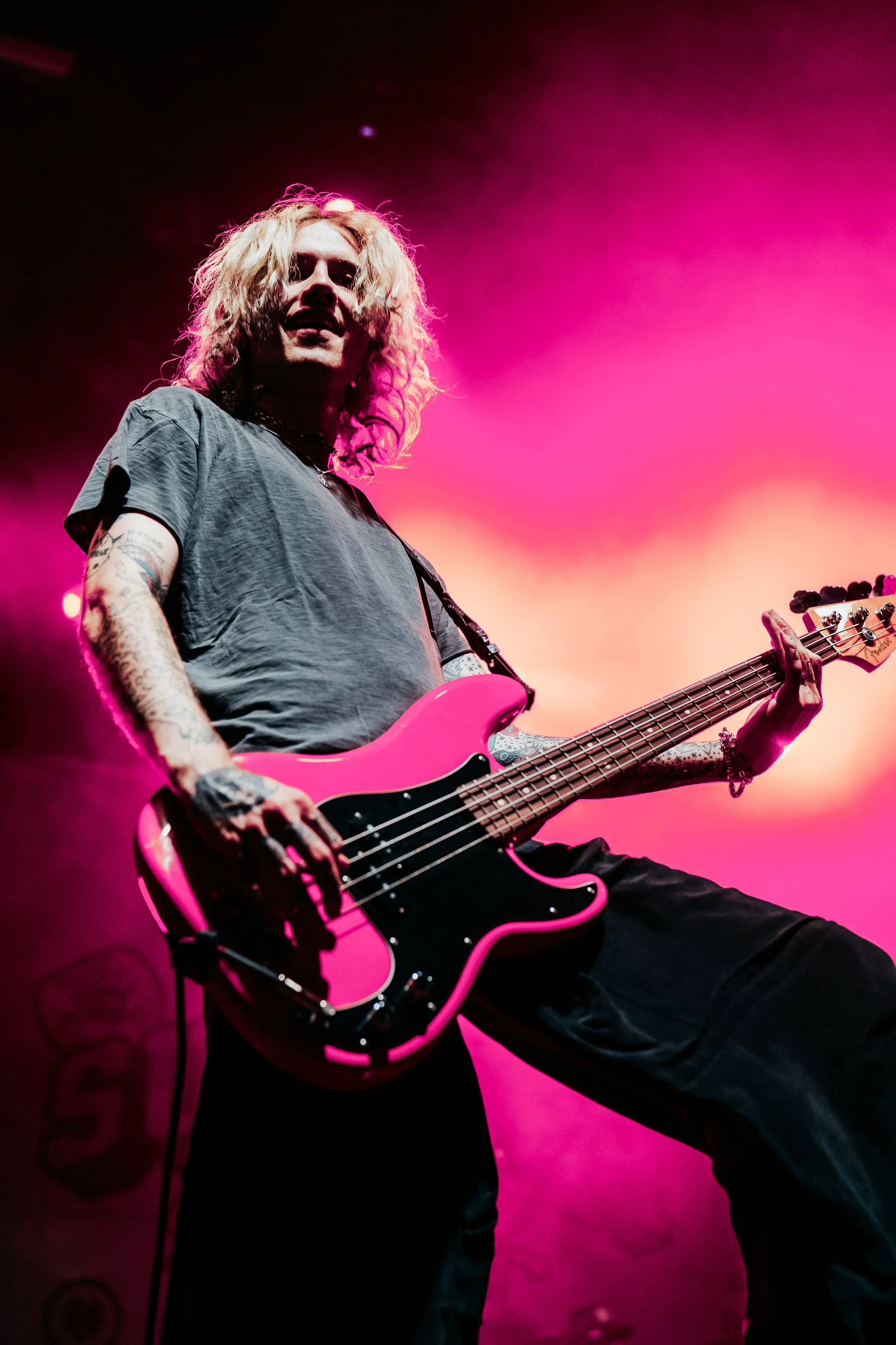 A musician playing a pink bass guitar on stage with pink and orange lighting.