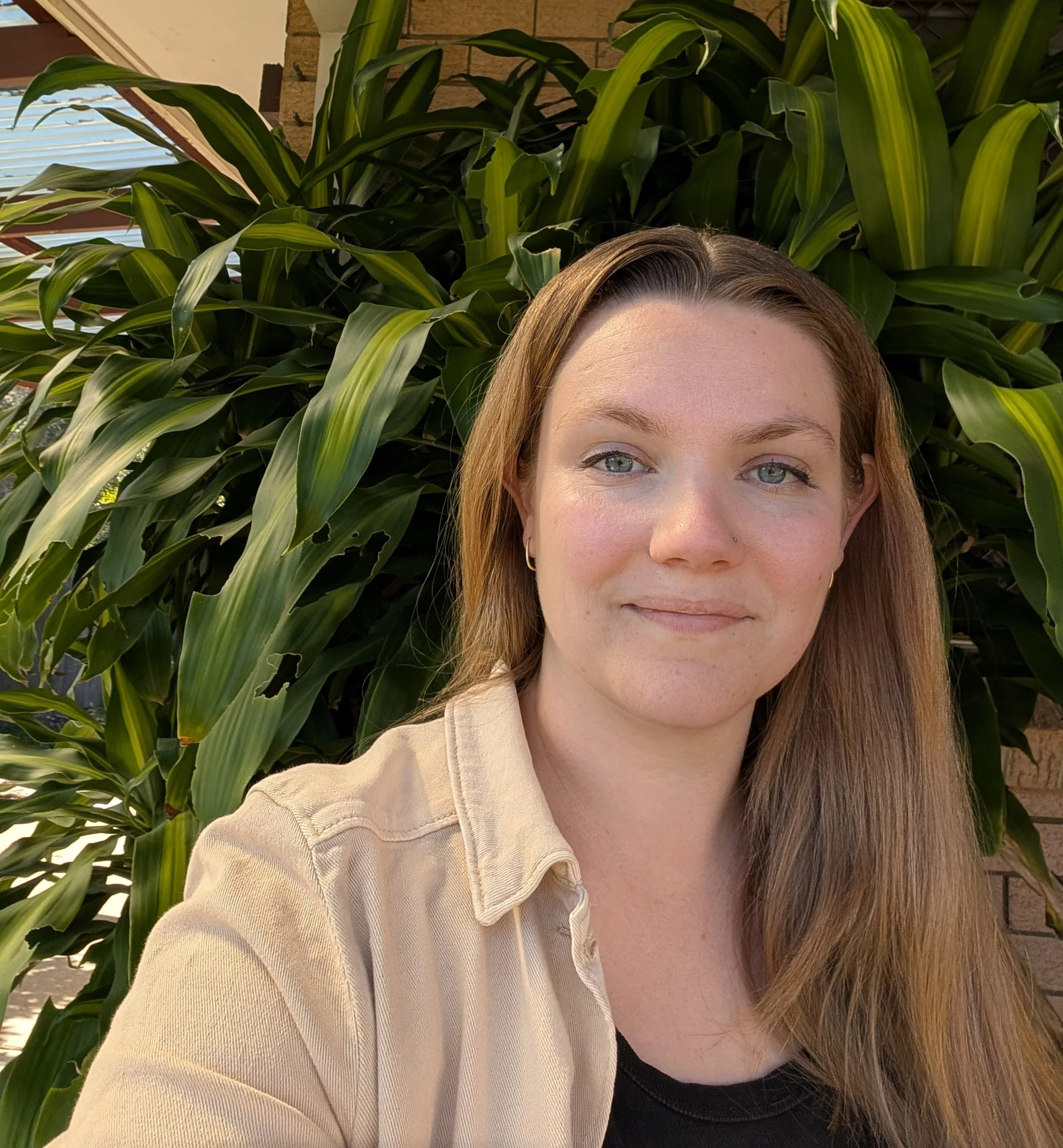 Close-up of a woman with brown hair and light makeup standing in front of large green plants with a brick wall and window with blinds in the background.