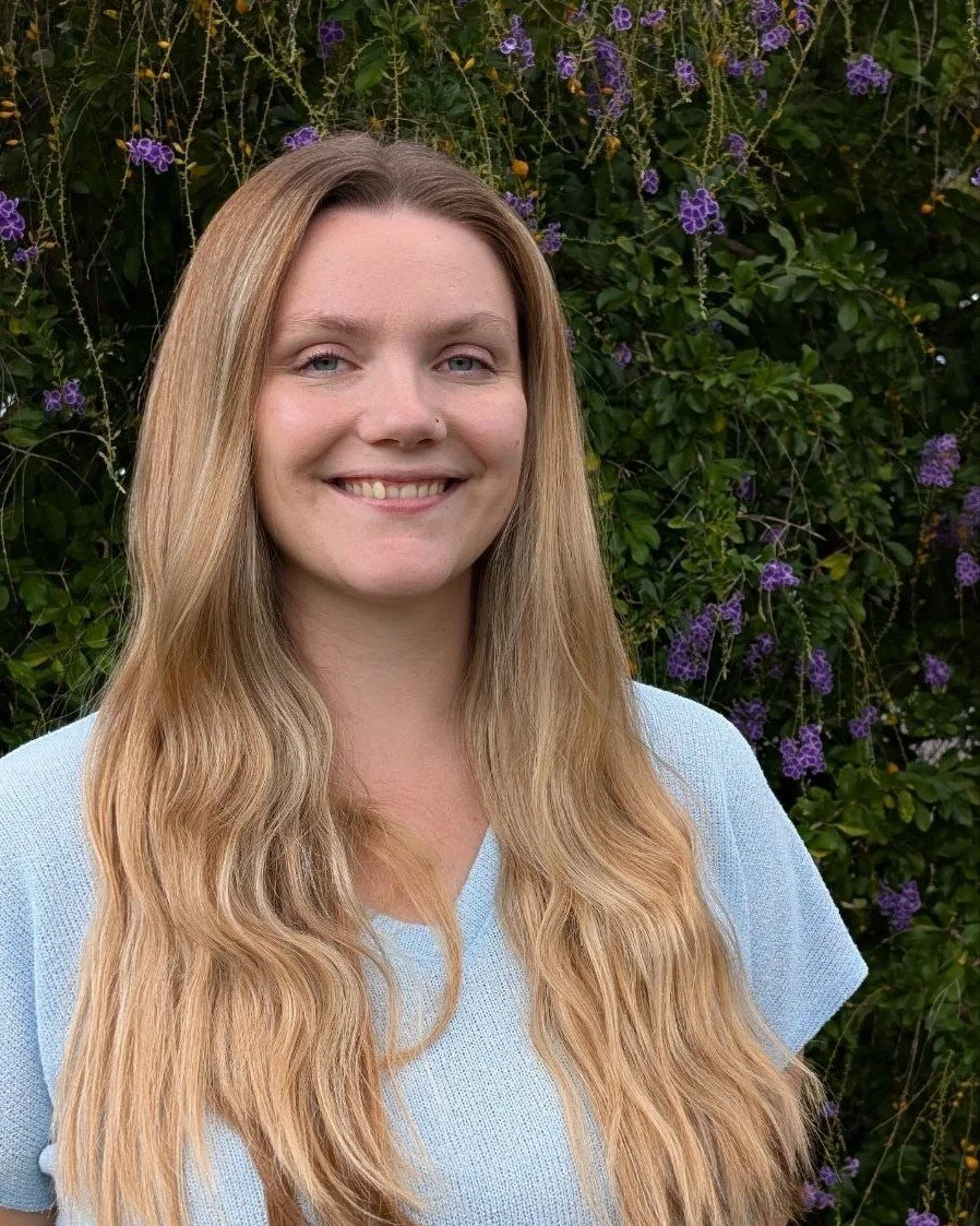 A woman with long, wavy blonde hair smiling outdoors in front of a bush with purple flowers.