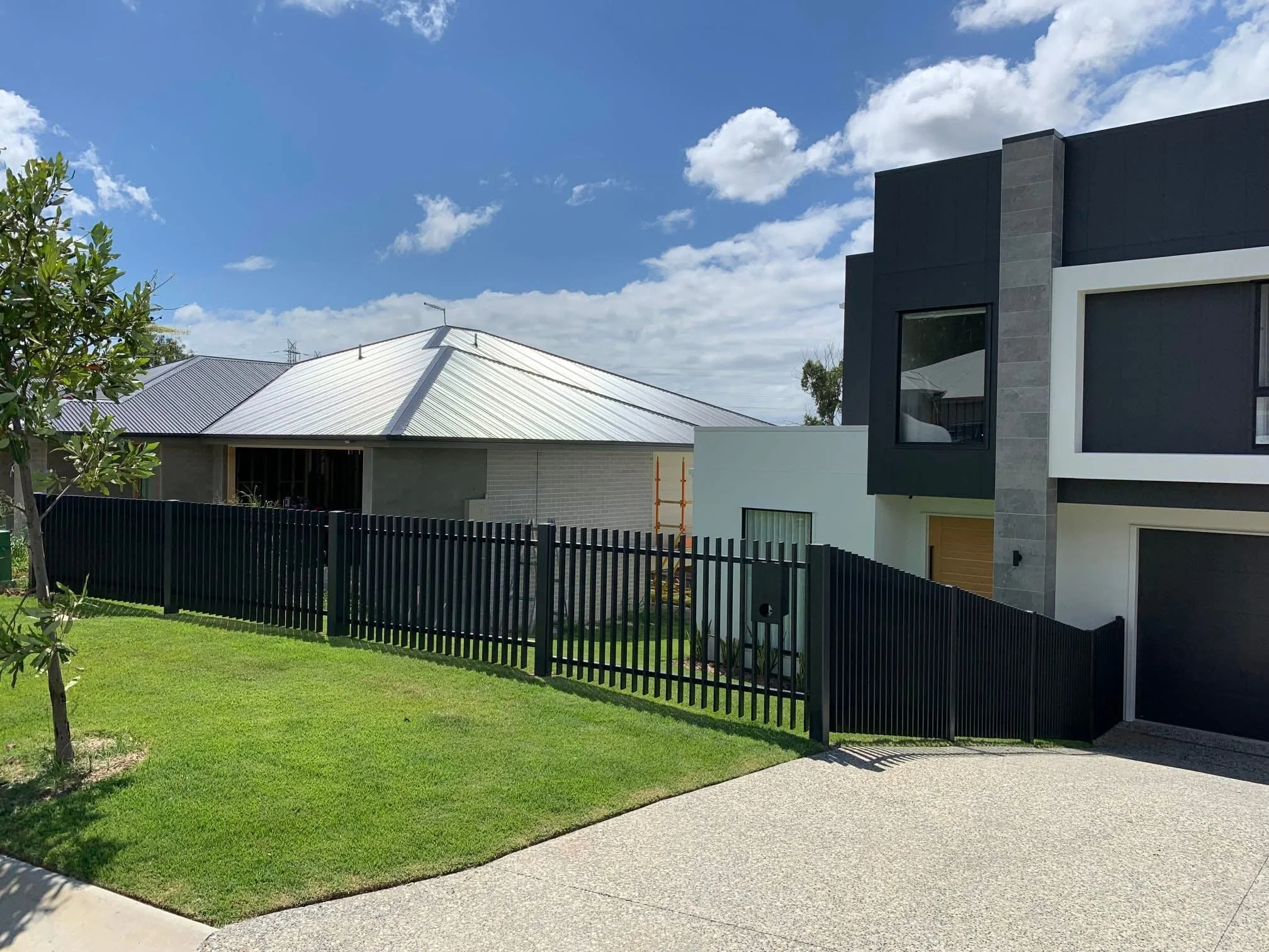 Modern two-story house with black and gray exterior, large windows, and a black fence along the front yard with green grass and a concrete driveway, under a partly cloudy sky.