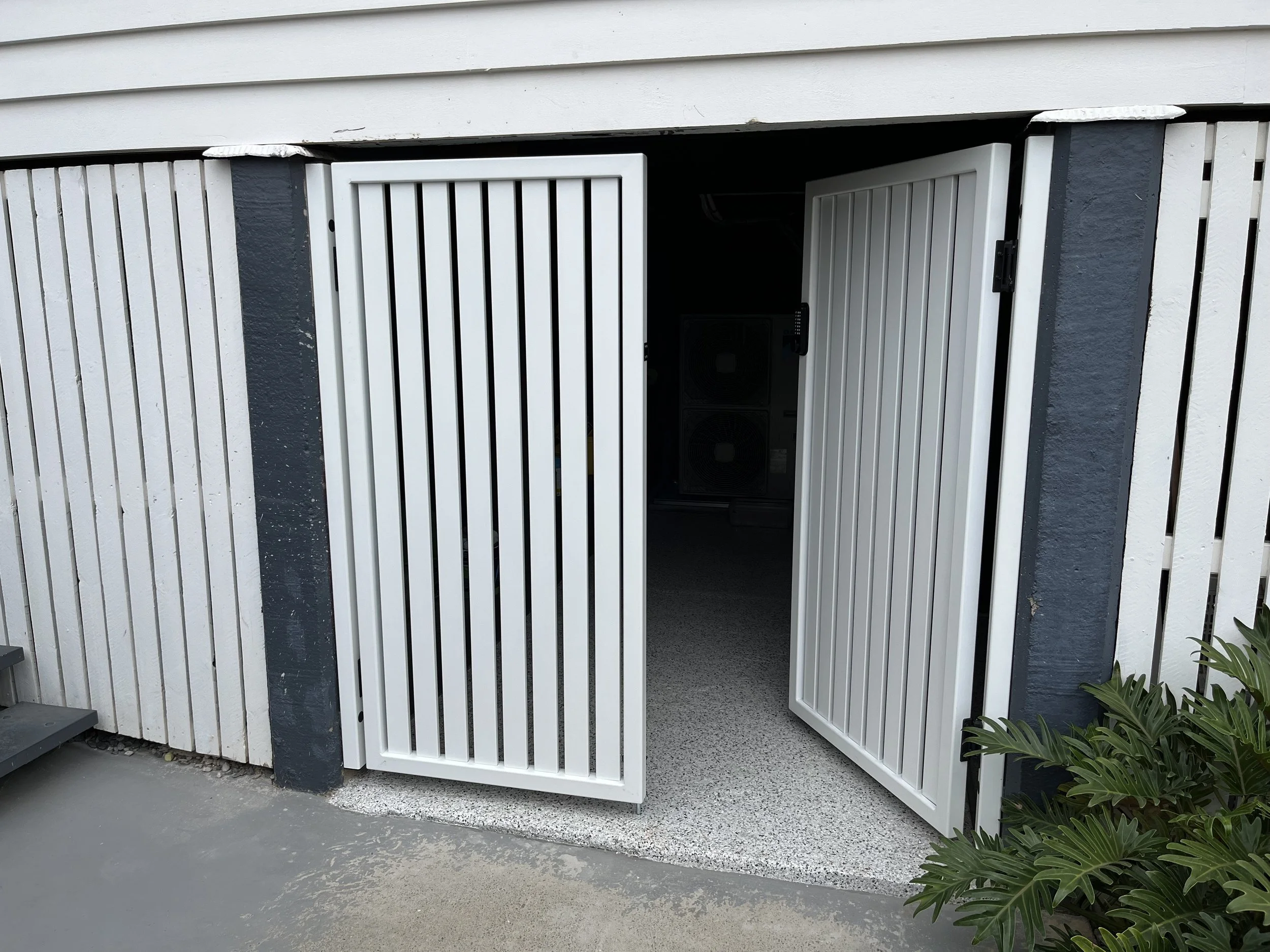 Exterior view of partially opened white metal gate, adjacent to a gray and white wooden wall and some green plants.