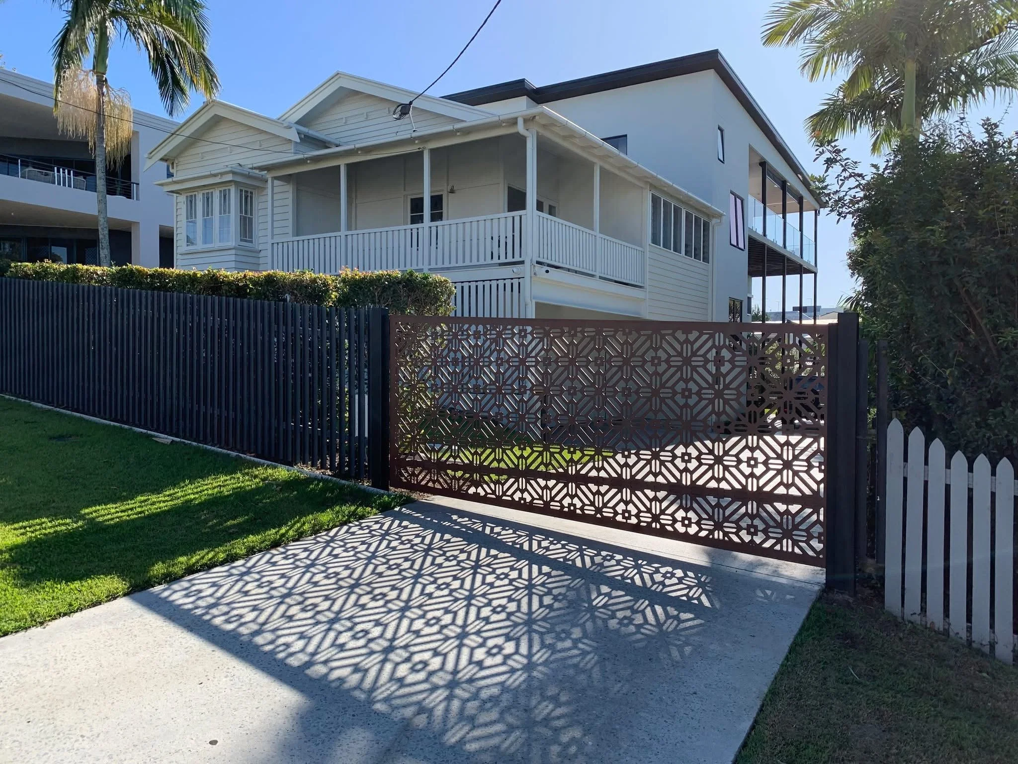 A white two-story house with balconies and a decorative metal gate casting shadows on the sidewalk, surrounded by palm trees and greenery.