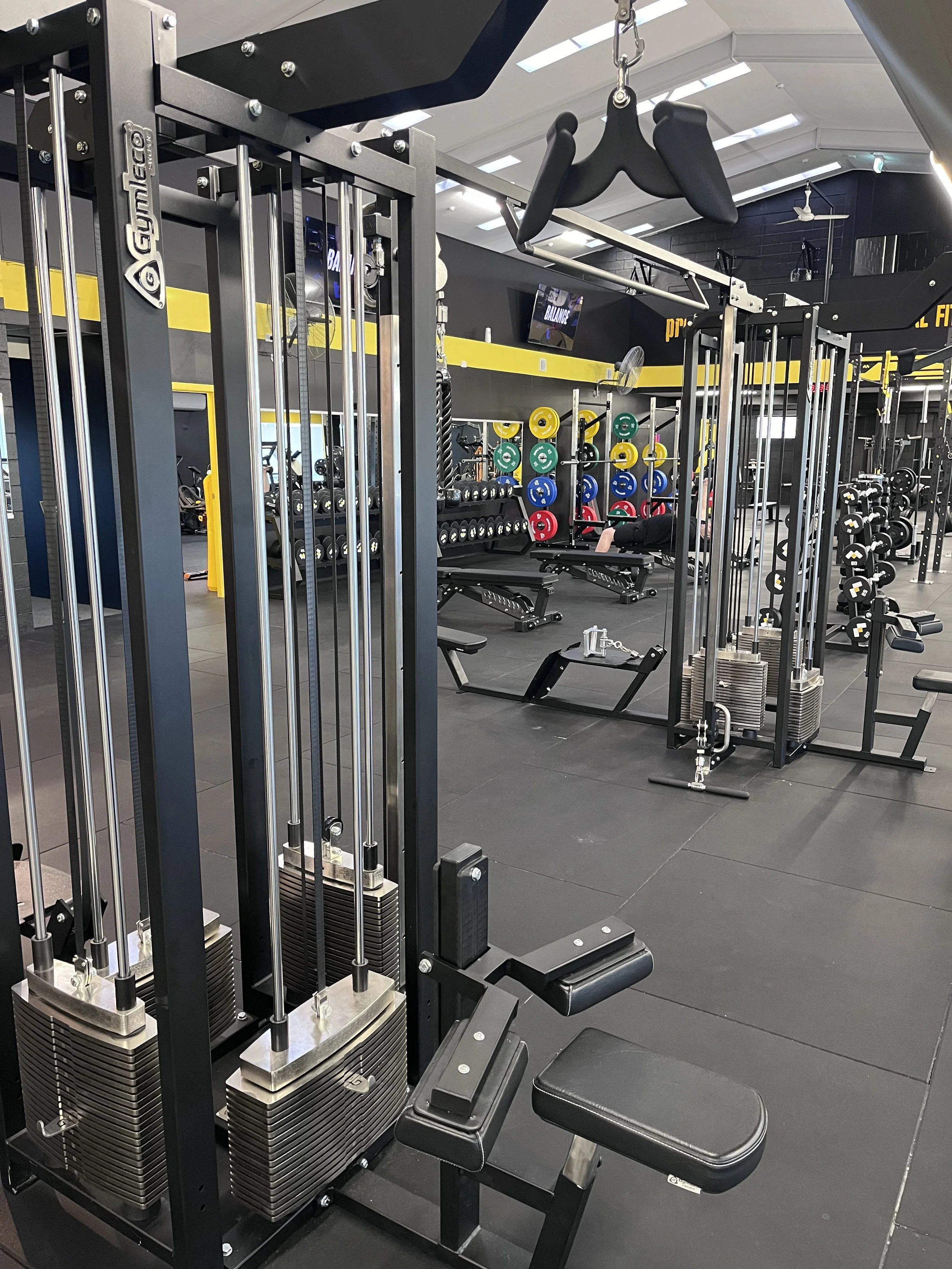Interior of a gym with weight training equipment, dumbbells, and weight plates, with a person exercising in the background.