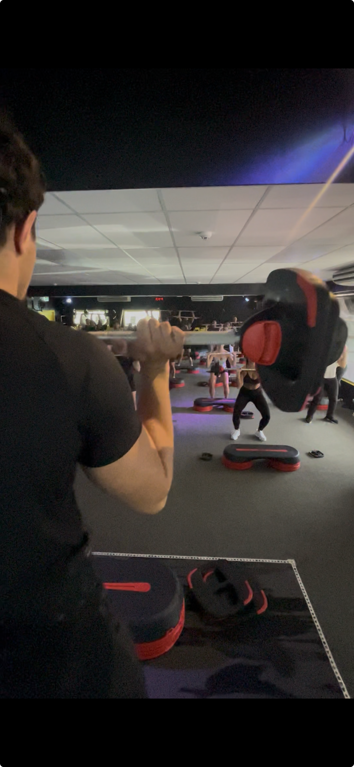 Person lifting a weighted barbell during a group fitness class in a gym with participants exercising in the background.