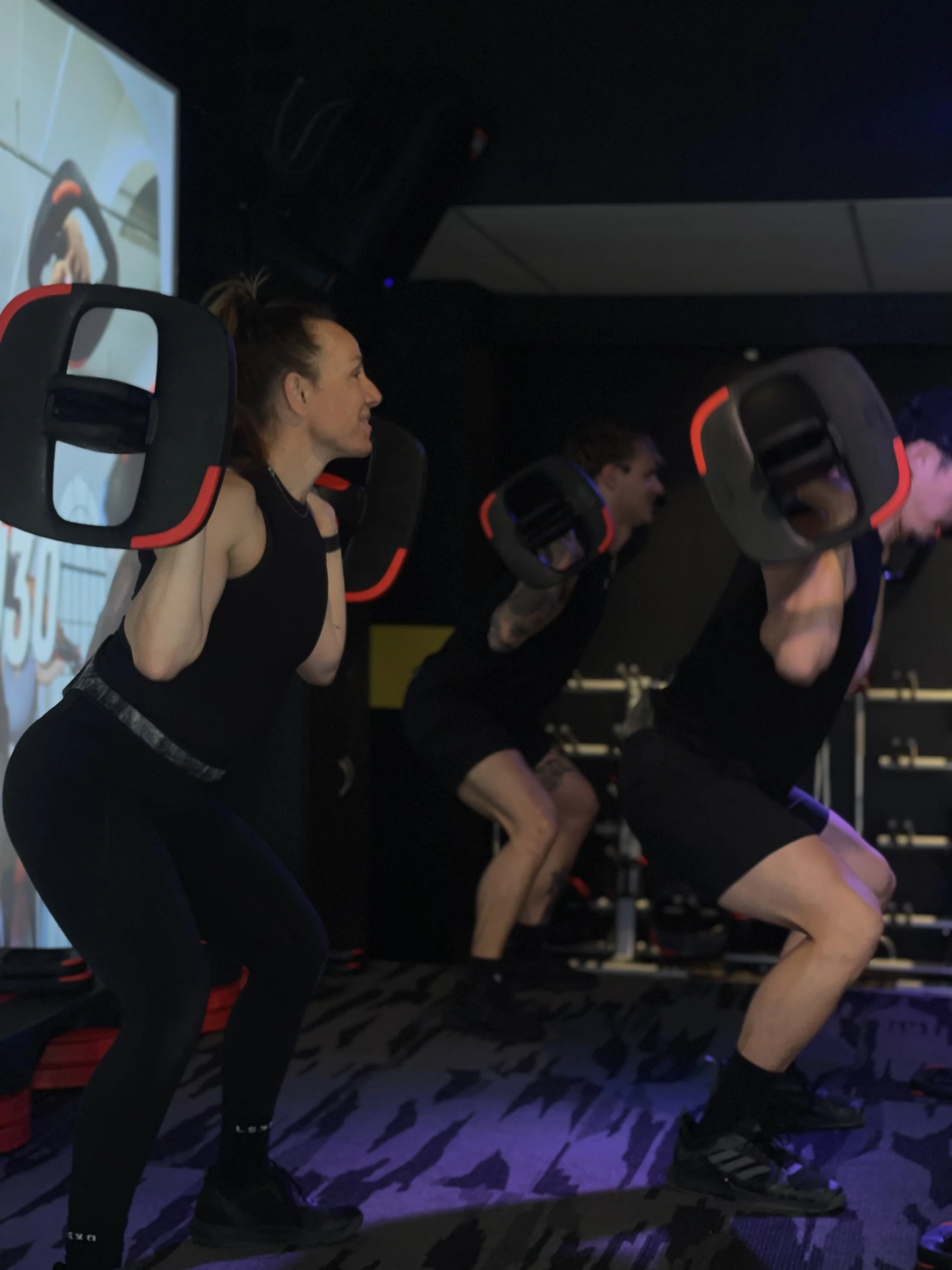 Group of people participating in a fitness class doing squats with large weighted pads on their shoulders in a dimly lit gym.