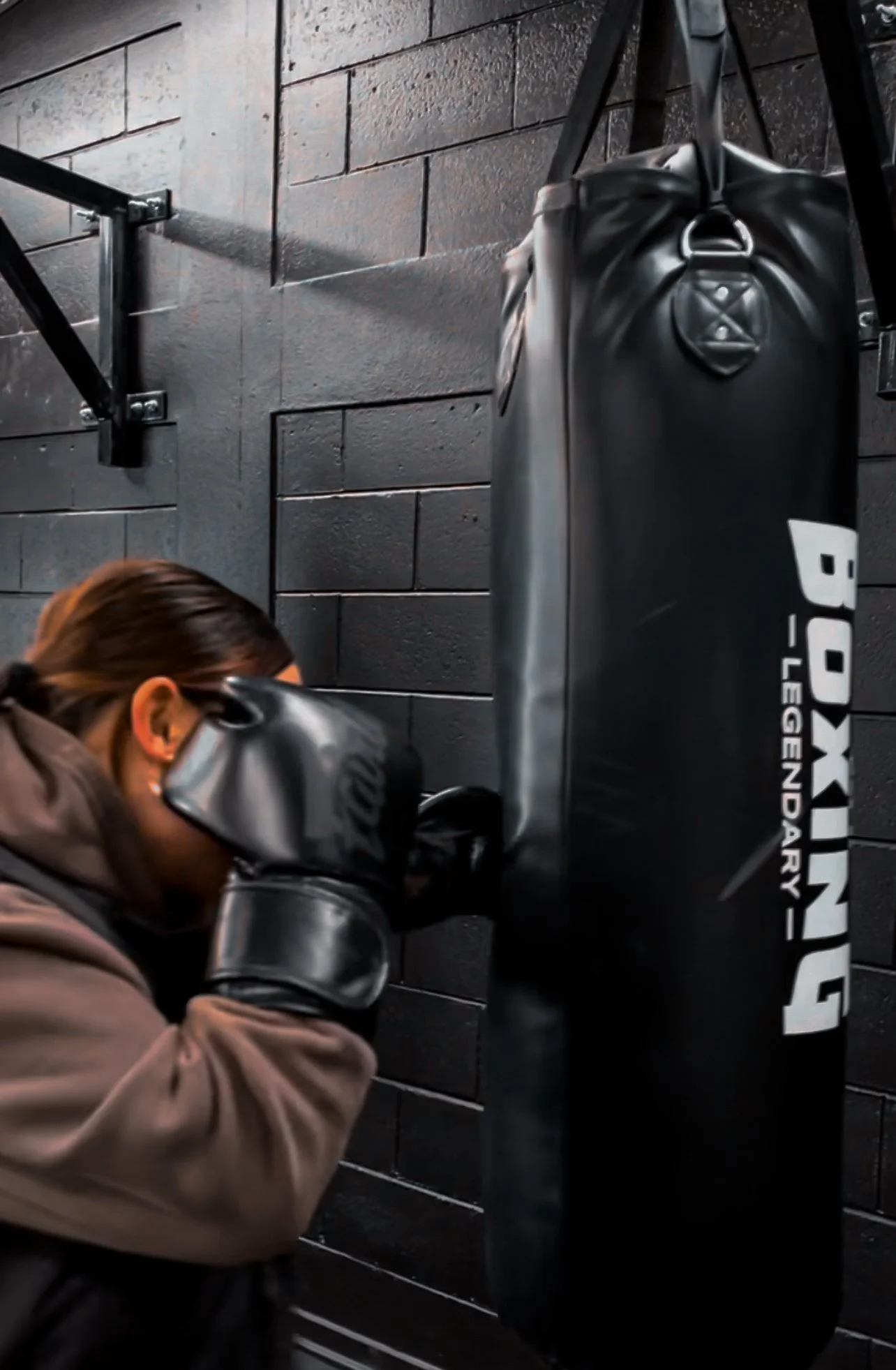 Person wearing boxing gloves and brown hoodie hitting a black boxing punching bag in a gym with black brick walls.