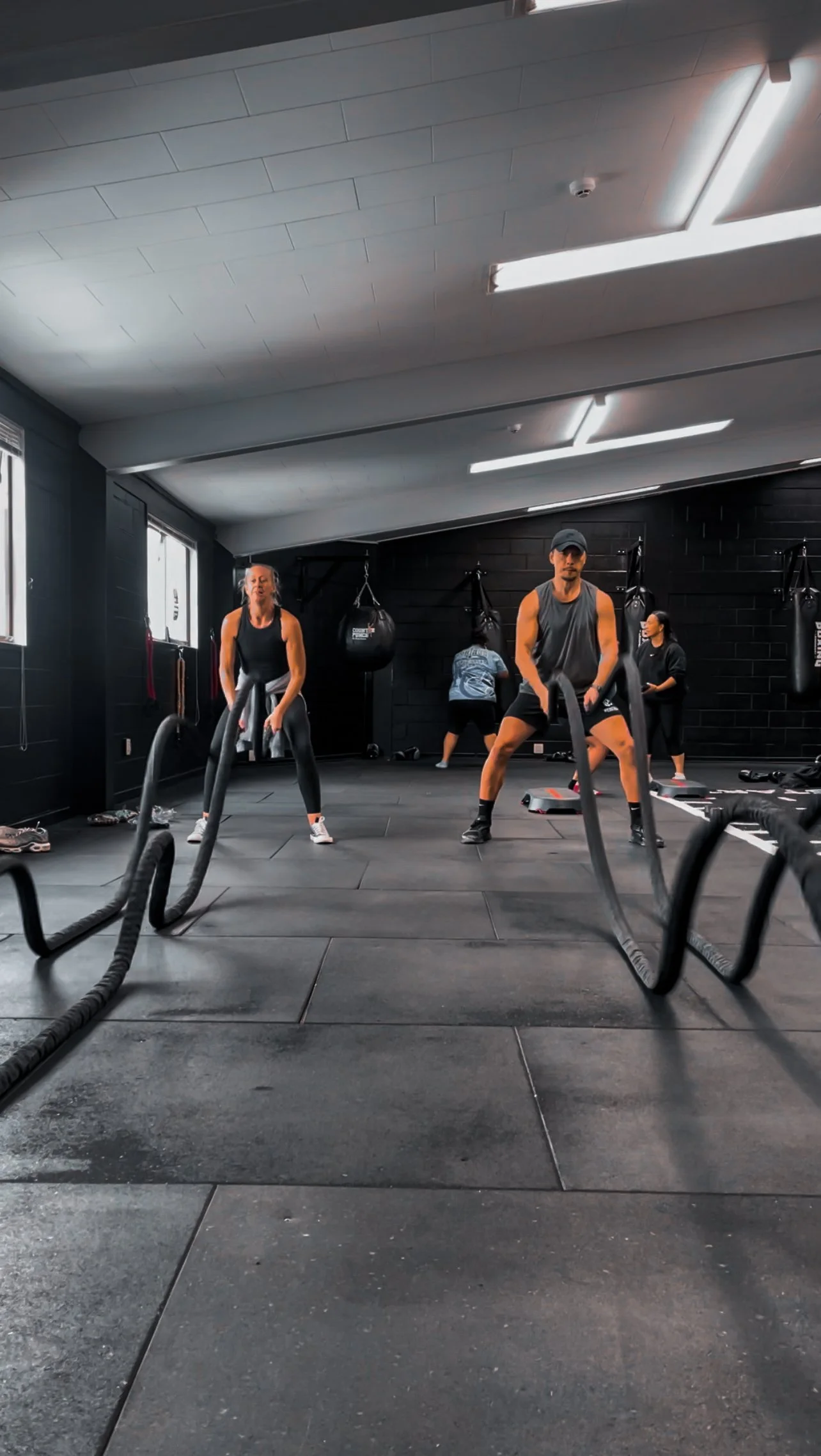 People doing a battle rope workout in a gym. Multiple individuals are engaged in various exercises, with some in the background near punching bags.