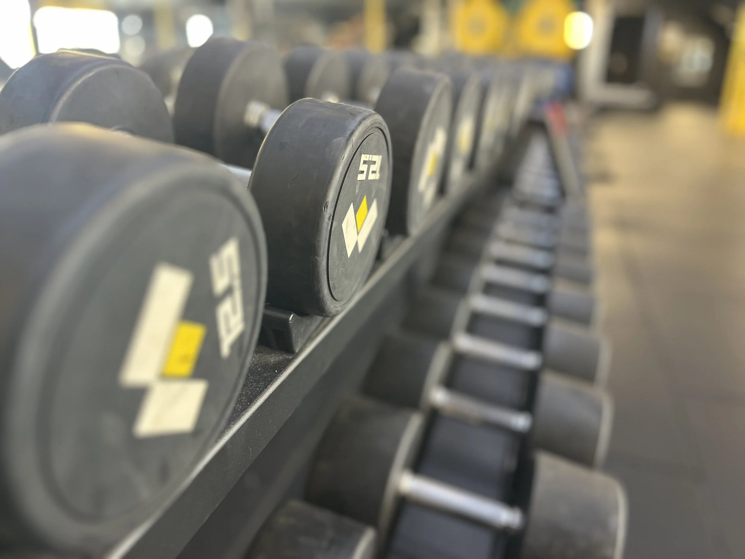 A row of black dumbbells with yellow and white markings on a rack in a gym, slightly out of focus.