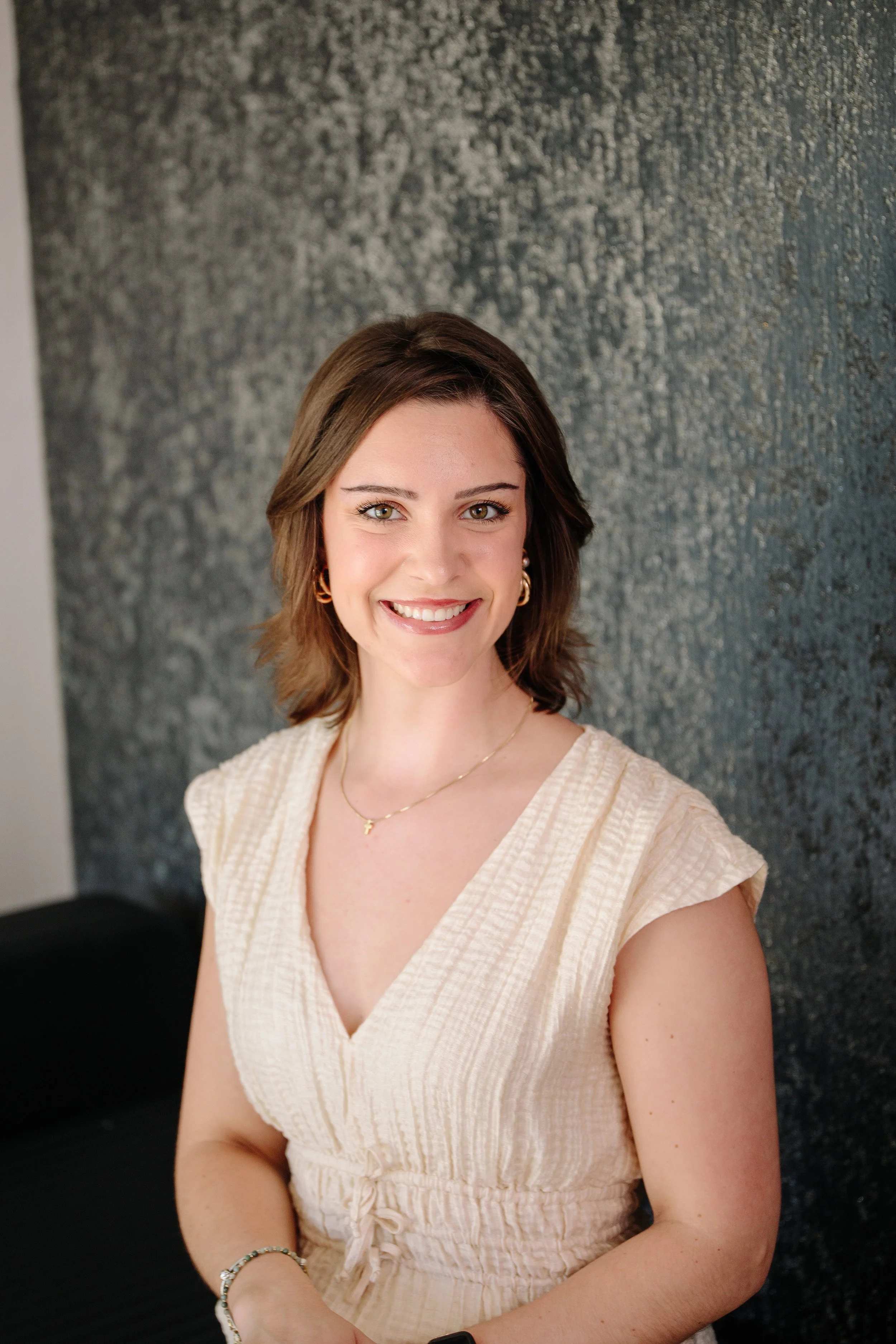A woman with shoulder-length brown hair, smiling and wearing a beige top, jewelry, and a watch, standing in front of a textured dark wall.