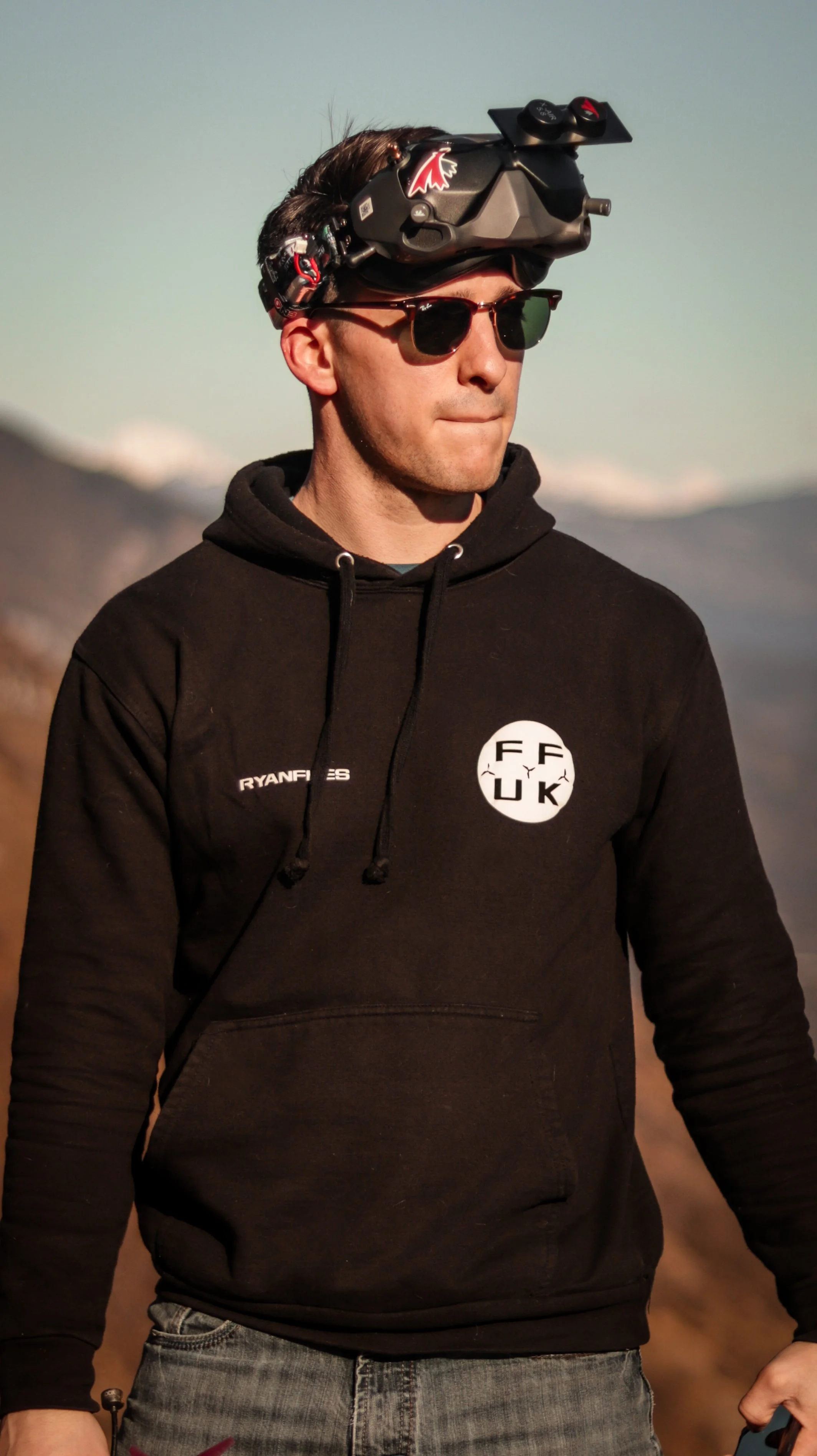 A man wearing sunglasses, a black hoodie with a circular logo reading 'FF UK', and a head-mounted drone controller with goggles, standing outdoors with mountains in the background.
