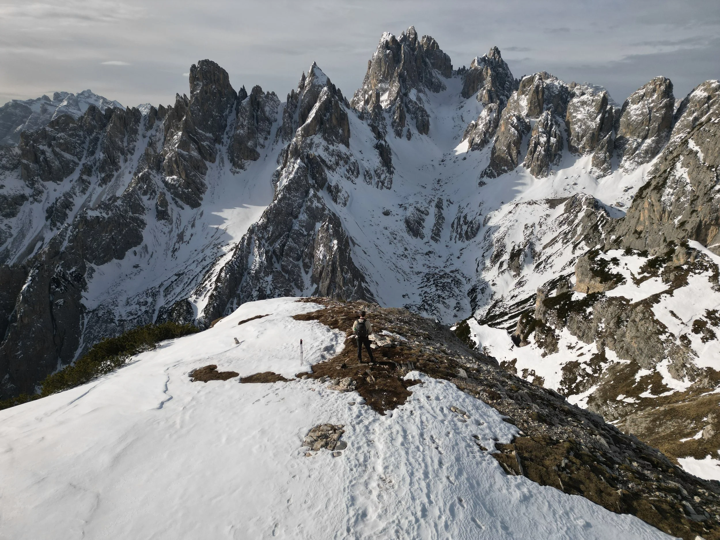 A hiker standing on a snowy mountain ridge overlooking a rugged snow-capped mountain range.