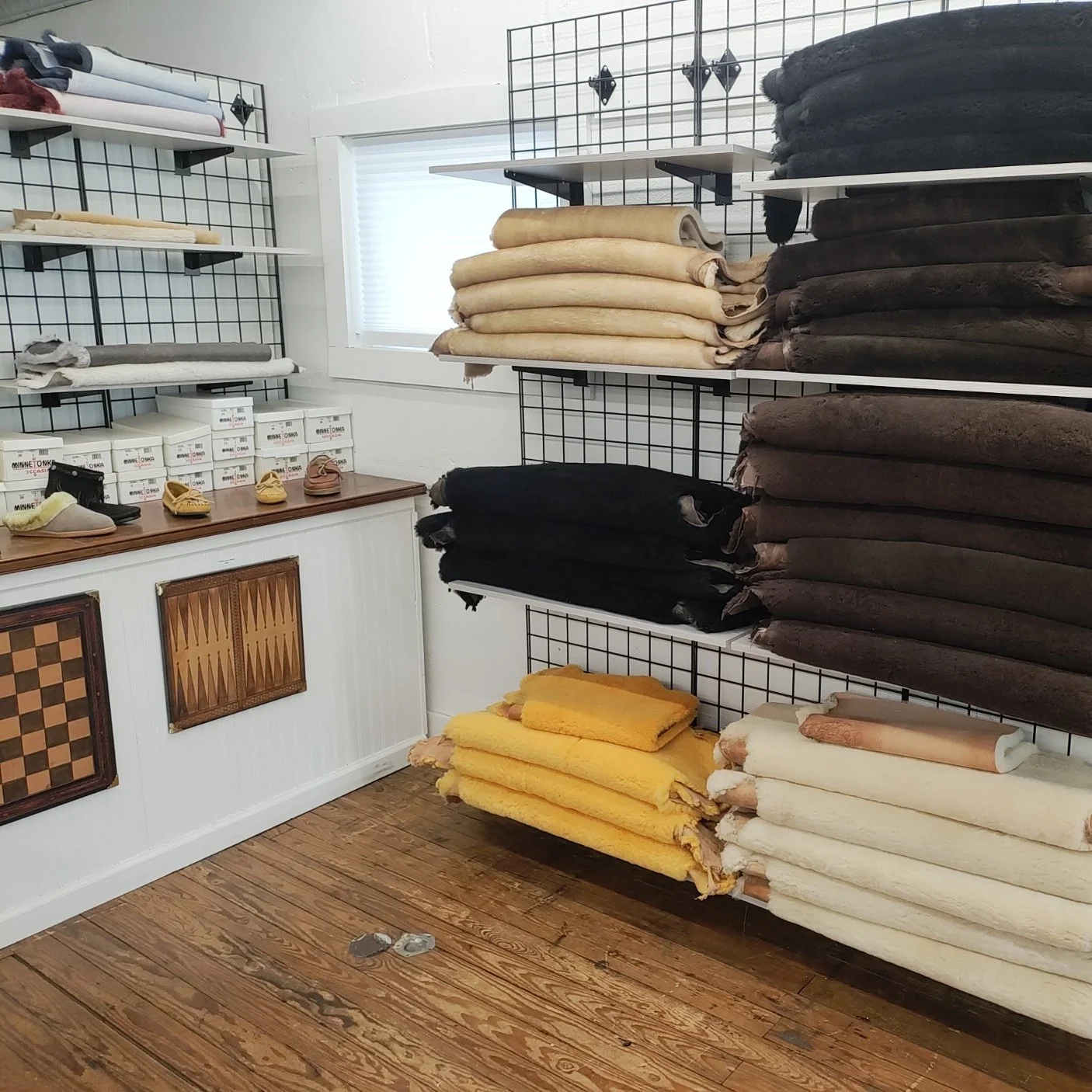 Display of various rolled and folded blankets and fleece throws in colors including cream, black, brown, yellow, and white, arranged on wire shelves in a store. Shoes are displayed on a wooden counter underneath the shelves, with boxed shoes stacked nearby. The room has a wooden floor and a window with blinds.