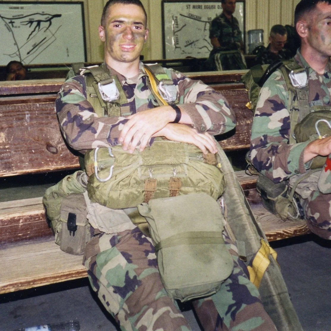 A soldier in a camouflage uniform sitting on a wooden bench with arms crossed, smiling at the camera, in a room with others in similar uniforms and maps on the wall.