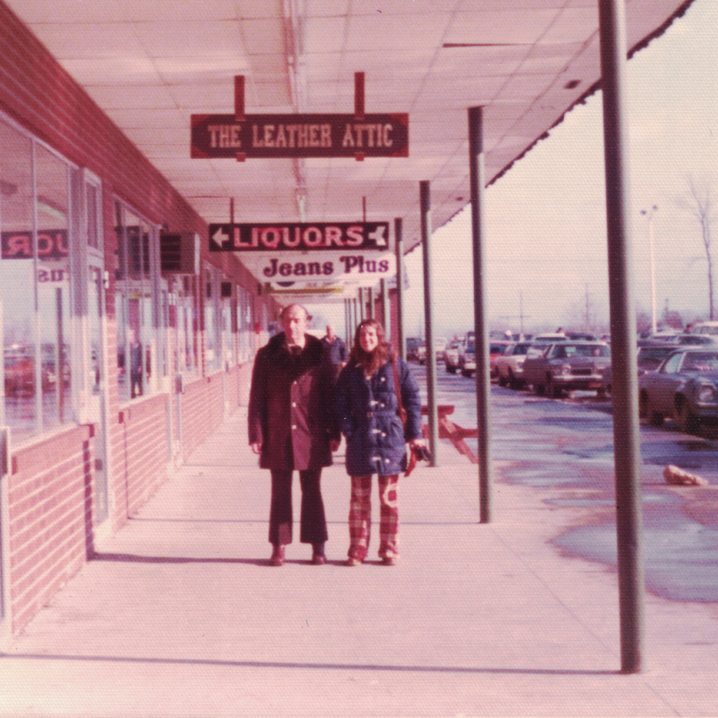 A shopping plaza with signs for a leather boutique, liquor store, and jeans store, with two women and a child walking on the sidewalk.