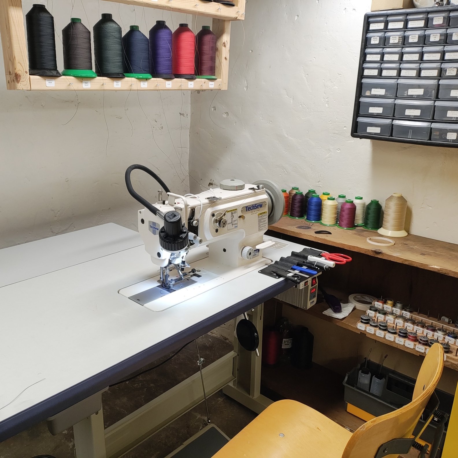 A workspace with a sewing machine, colorful thread spools, scissors, and organized storage bins. There are shelves with labeled small containers and spool displays, set in a room with white, textured walls and a wooden work surface.