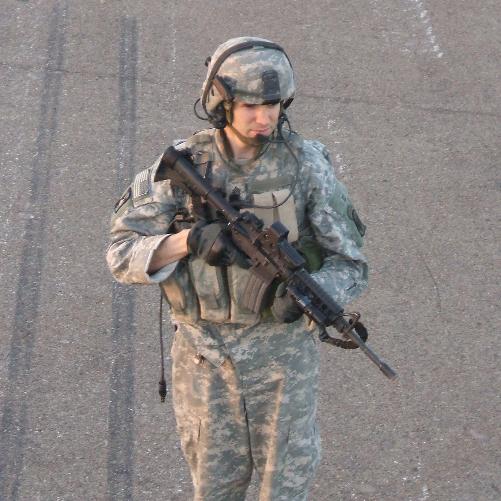 Military soldier wearing camouflage uniform and helmet, holding an assault rifle, standing on asphalt surface.