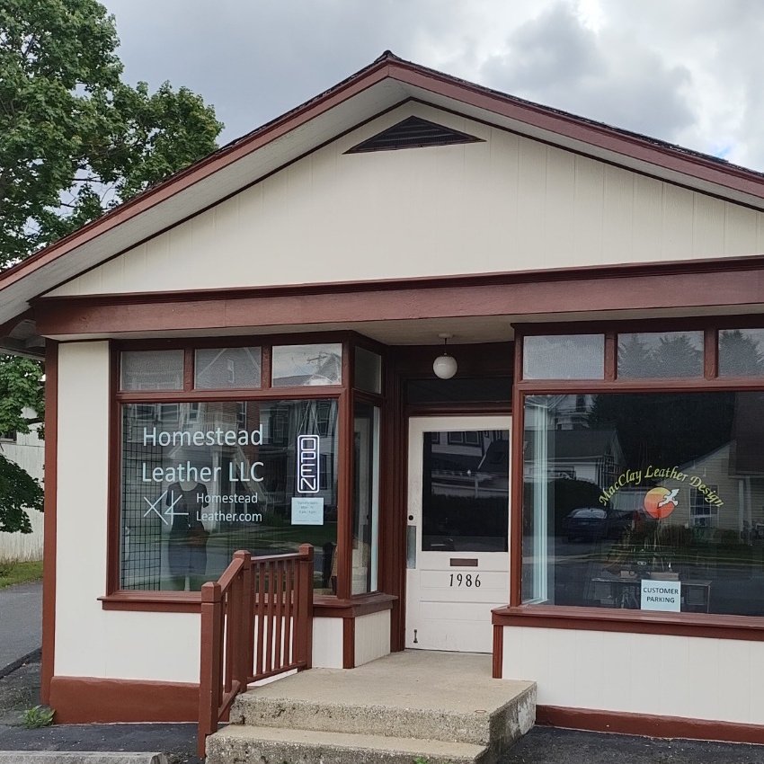 Exterior of a small storefront with a sign reading Homestead Leather LLC, with a glass window and door numbered 1986, a small ramp, and references to leather products inside.