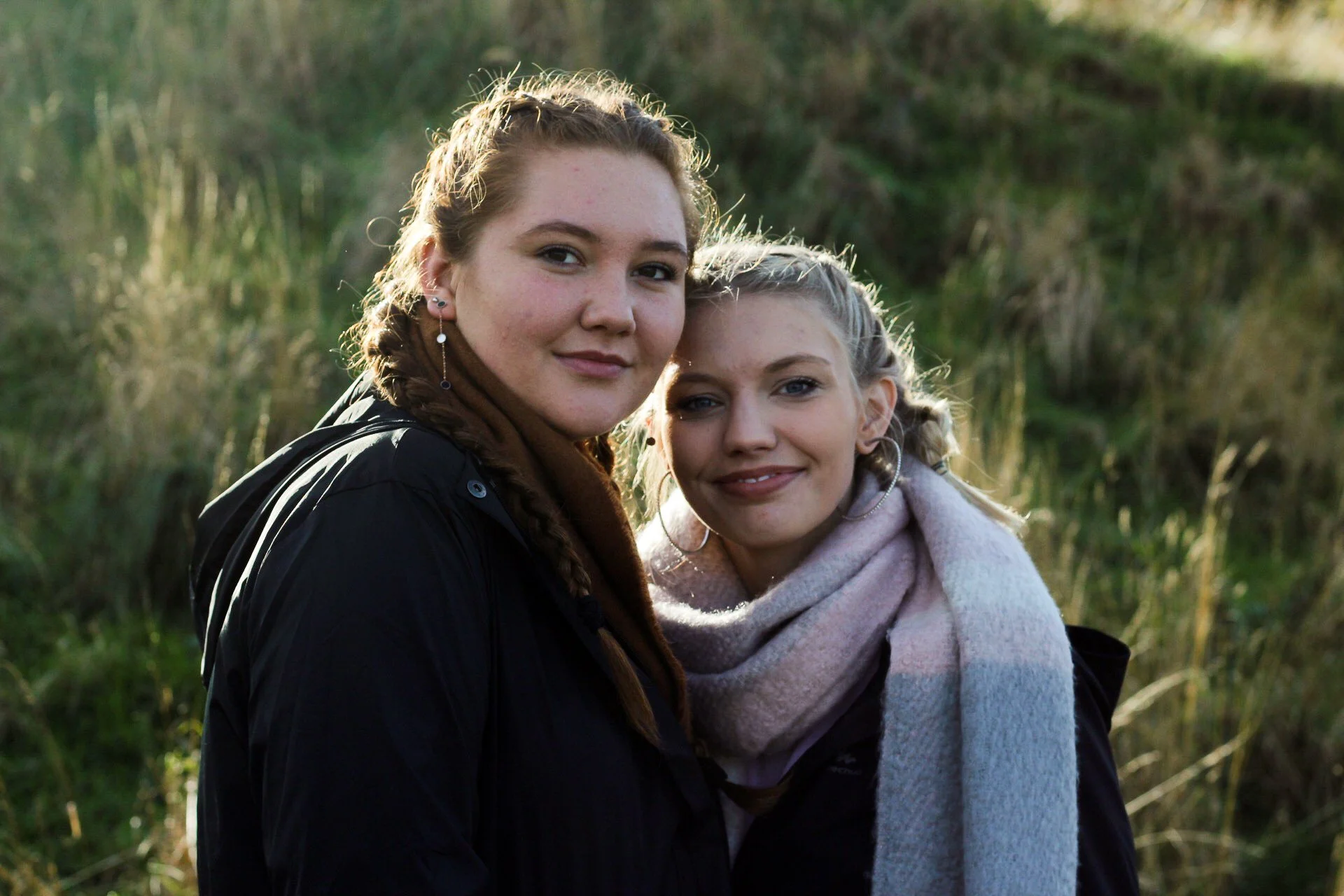 Two young women outdoors, standing close together, smiling, with natural greenery in the background.