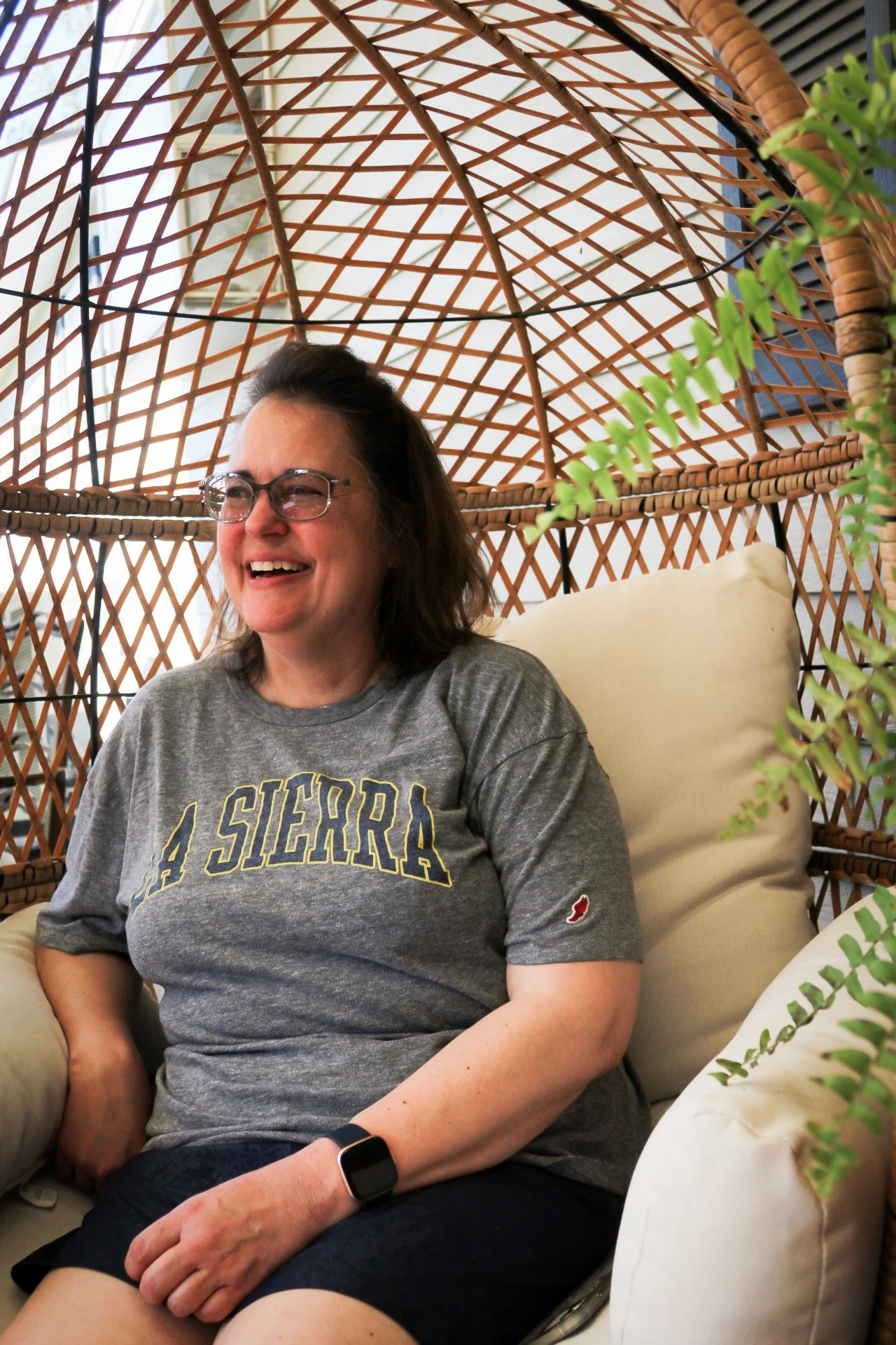 A woman sitting in a woven outdoor chair, smiling, wearing glasses, a gray T-shirt that says 'Sierra', and a smartwatch, with green plants around her.