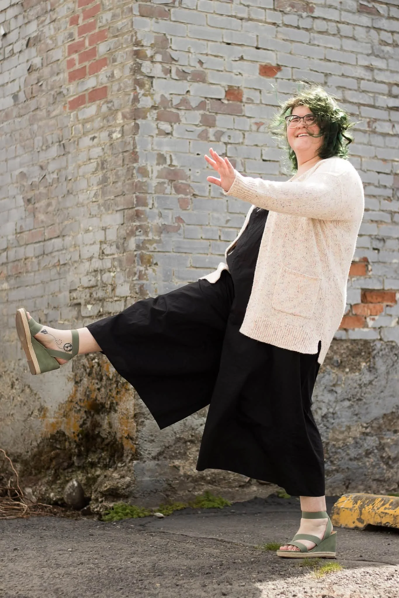 A woman with glasses and curly hair dressed in a black jumpsuit, light-colored cardigan, and green wedge sandals is posing outdoors against a brick wall, lifting one leg and smiling.