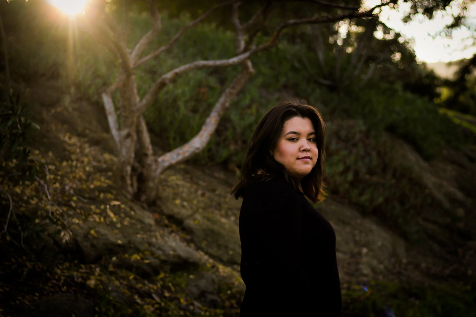 A woman with dark brown hair and a black top stands outdoors near a tree during sunset, with sunlight filtering through the branches.