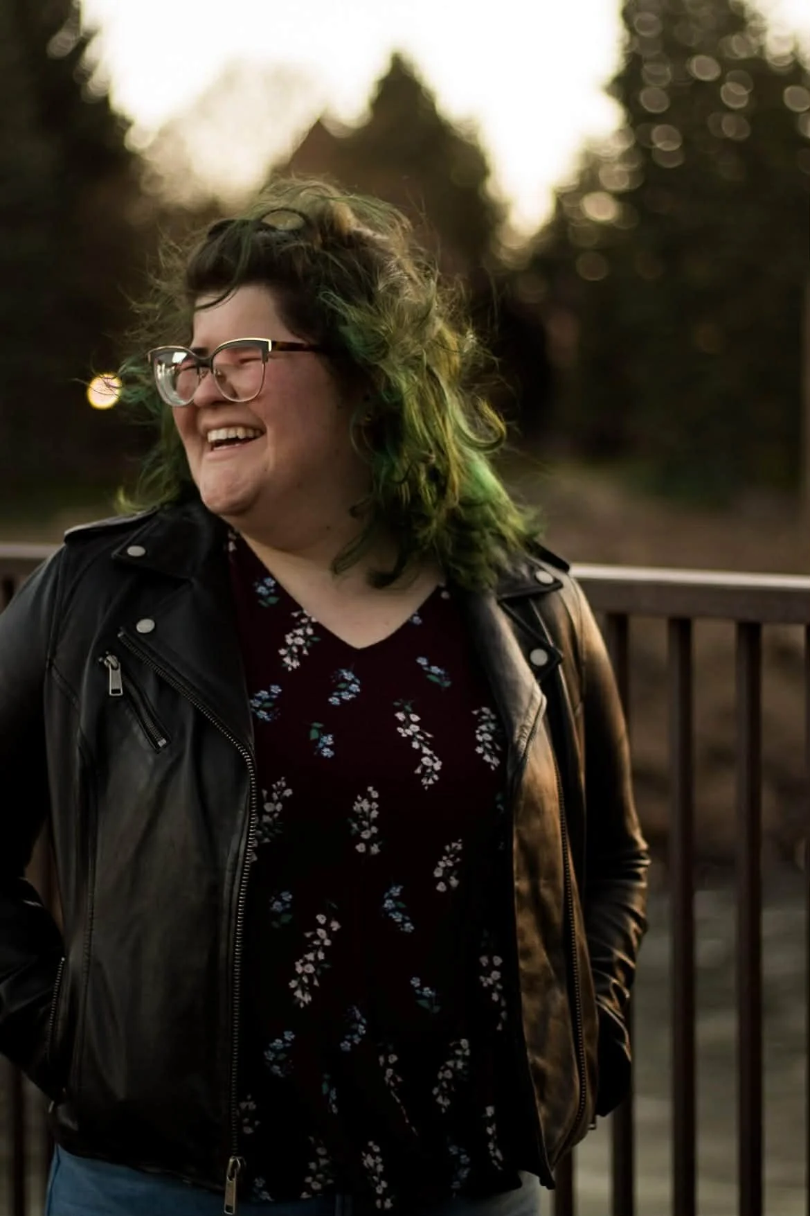 A person with short curly green-tinted hair and glasses, smiling and wearing a black leather jacket over a dark floral shirt, outdoors at sunset with trees and a fence in the background.