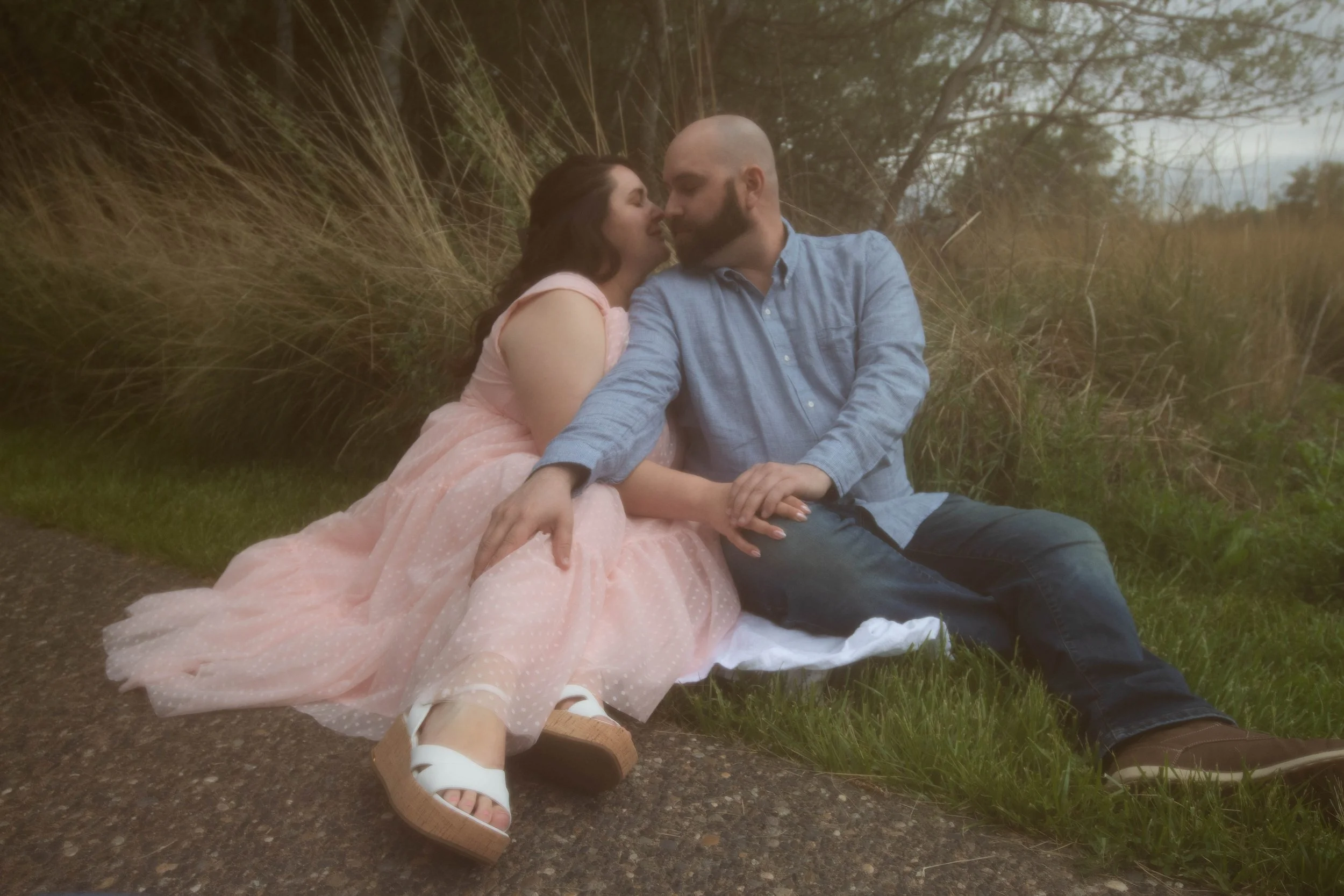 A couple sitting on the grass next to a walking path, leaning towards each other for a kiss, with trees and bushes in the background.