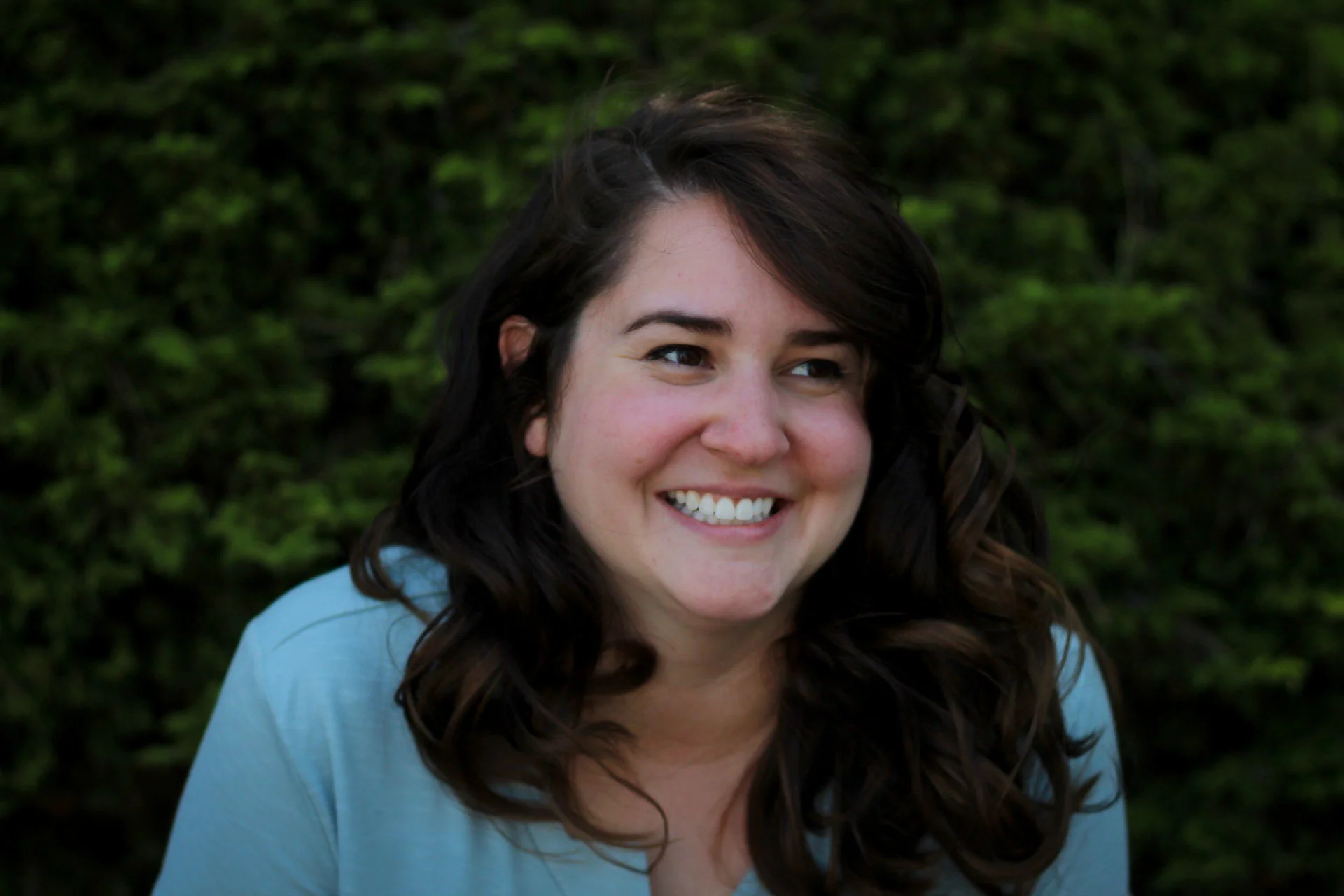 A smiling woman with dark, wavy hair wearing a light blue top stands outdoors with green foliage in the background.