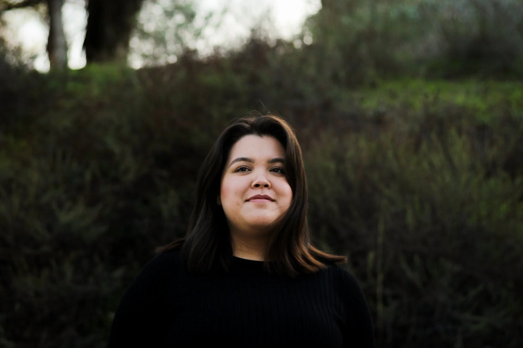 A young woman with dark hair and light skin, wearing a black sweater, standing outdoors in a natural setting with trees and green foliage in the background.