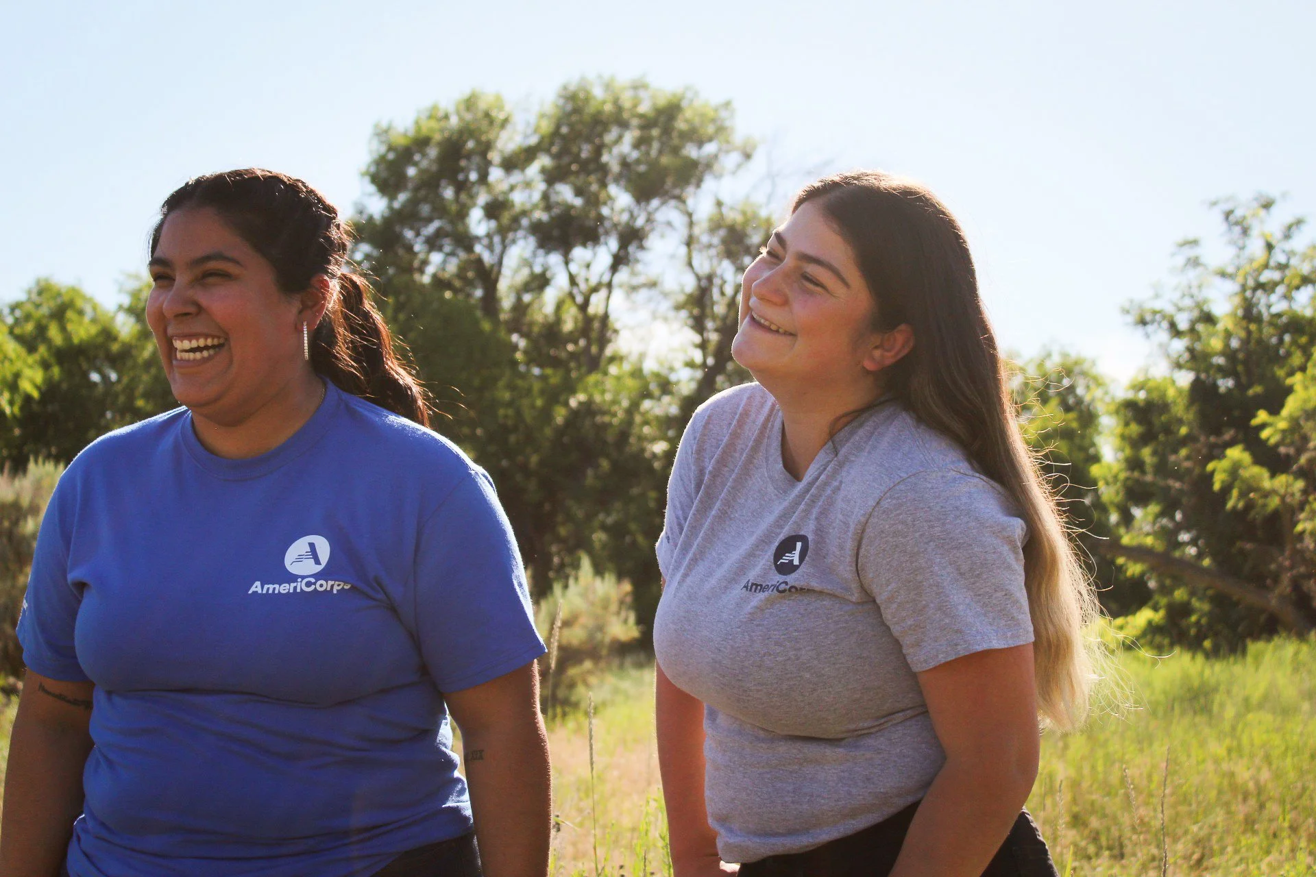 Two women wearing AmeriCorps shirts standing outdoors in a field, smiling and laughing with trees and a clear sky in the background.