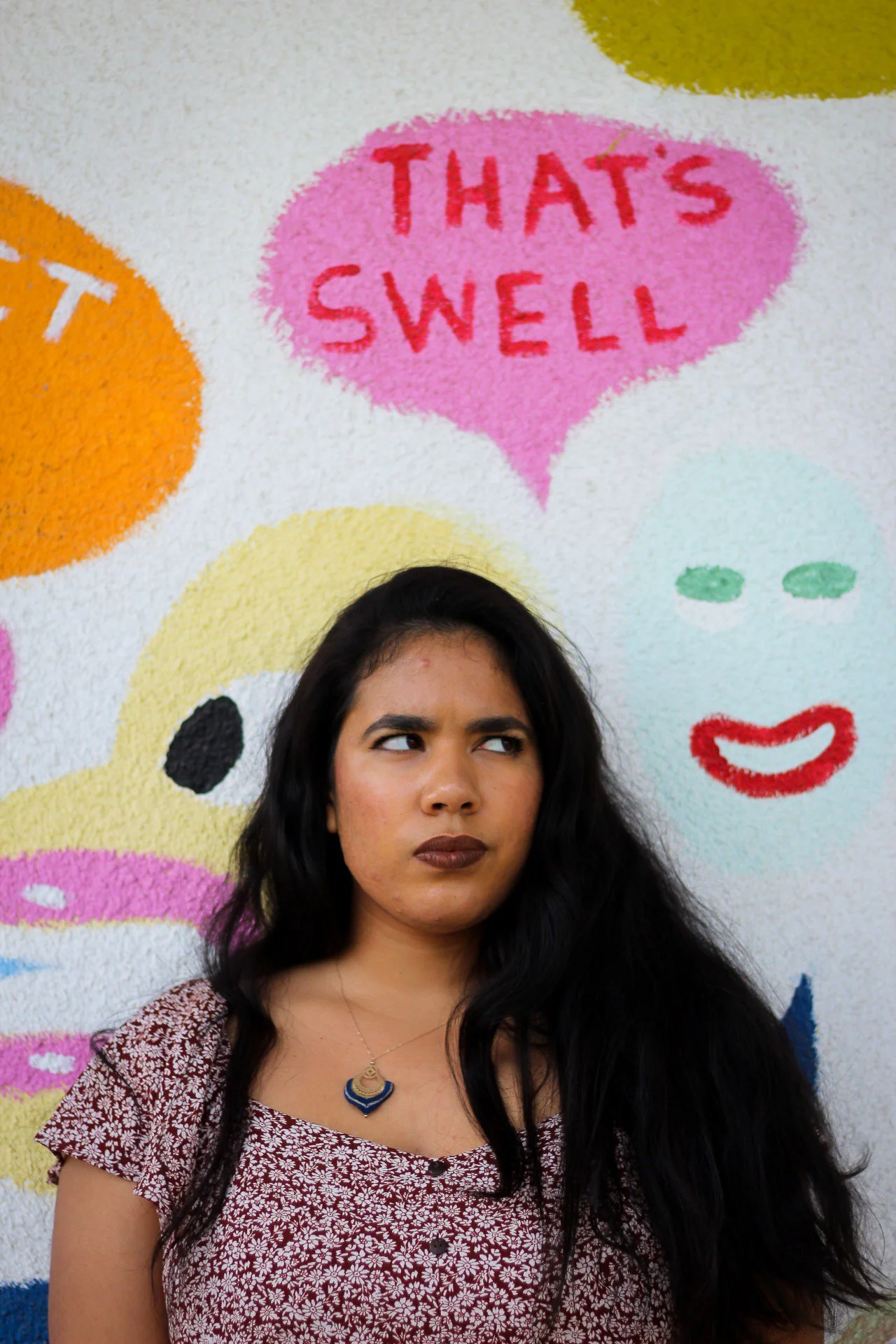 A woman with long dark hair looking to her left standing in front of a colorful mural with speech bubbles and abstract faces, one speech bubble says 'THAT'S SWELL' in red text on pink background.