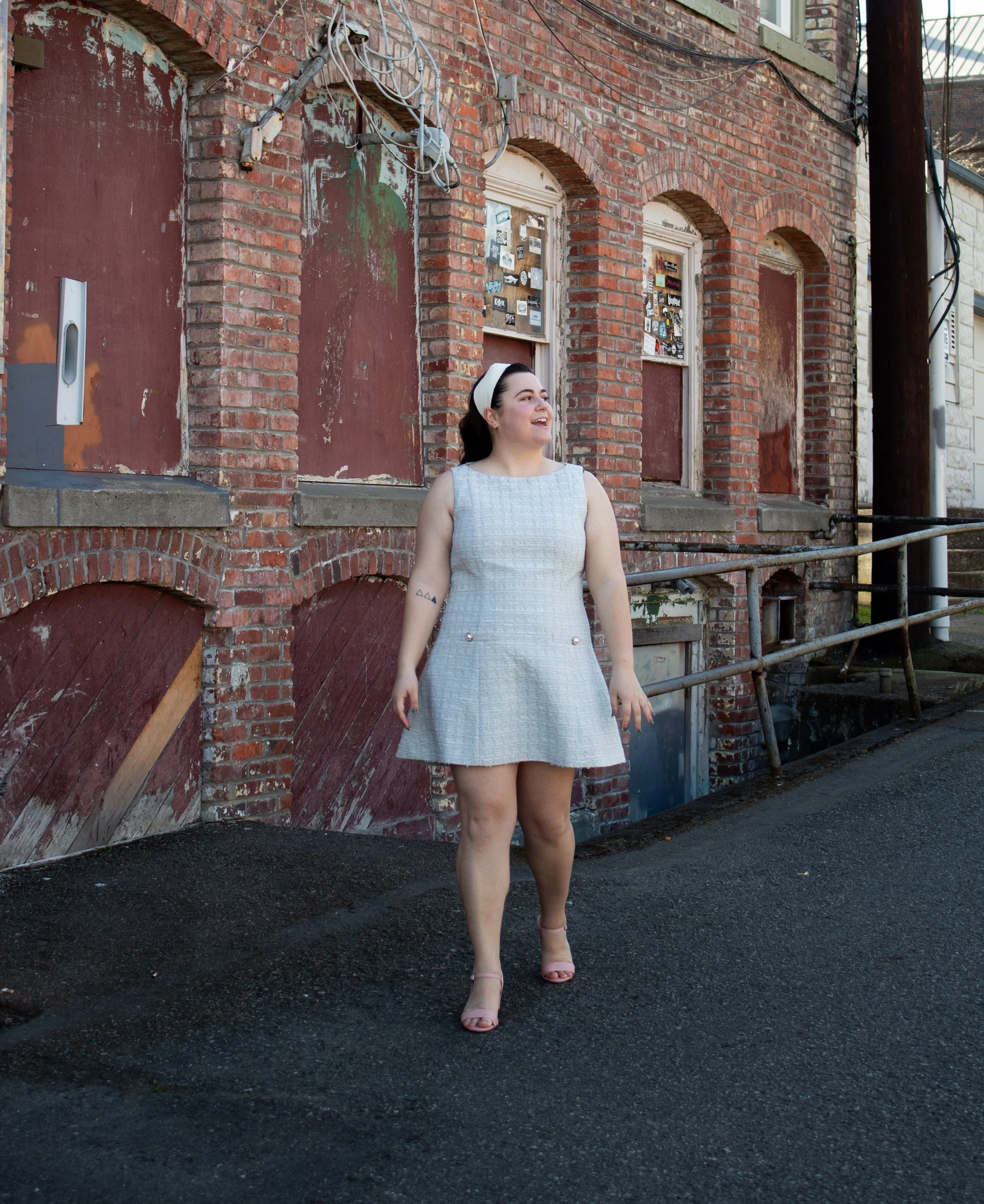 A woman in a sleeveless light gray dress and nude heels stands on a street in front of a weathered brick building with boarded-up windows, smiling and looking to the right.
